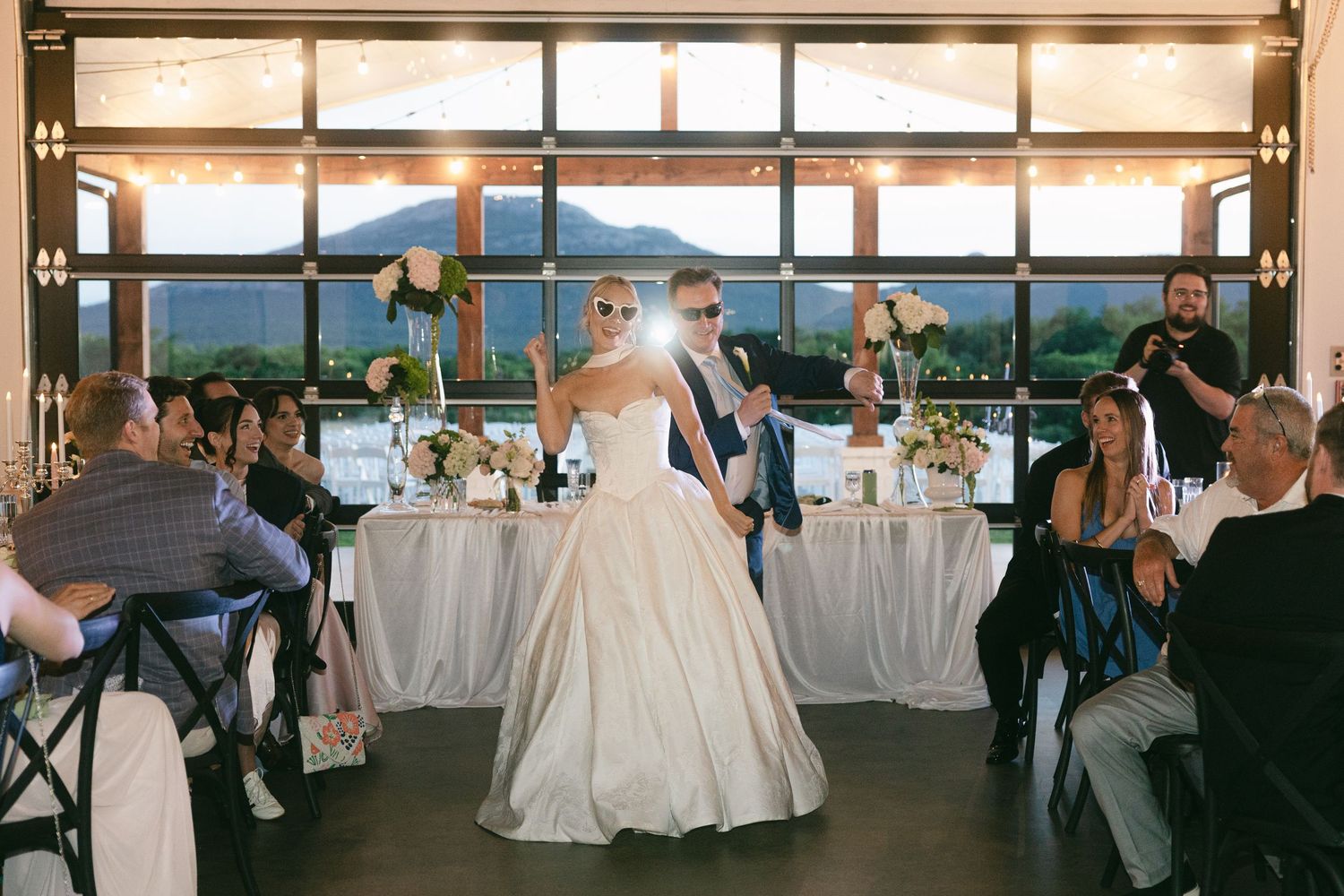 Bride and groom make grand entrance wearing sunglasses at elegant mountain view wedding reception.