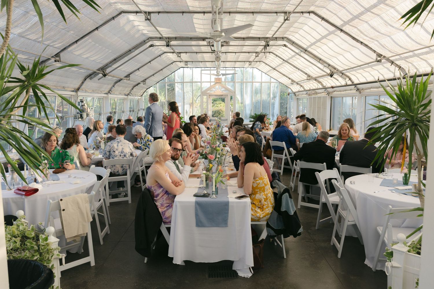 Elegant wedding reception in a glass greenhouse tent with white chairs, floral centerpieces, and lush greenery.