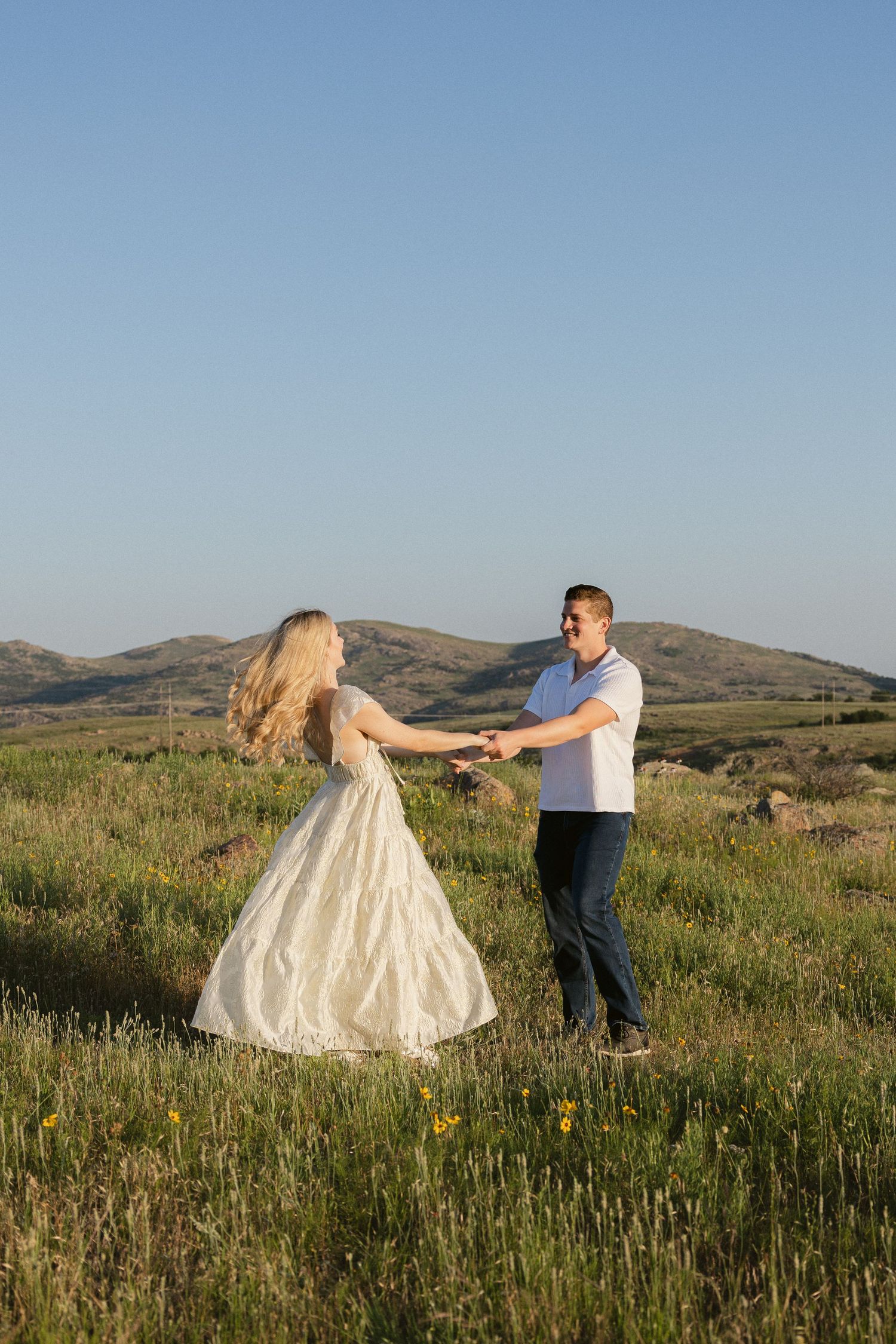 A couple dances together in a scenic meadow with wildflowers against a backdrop of rolling hills at sunset.