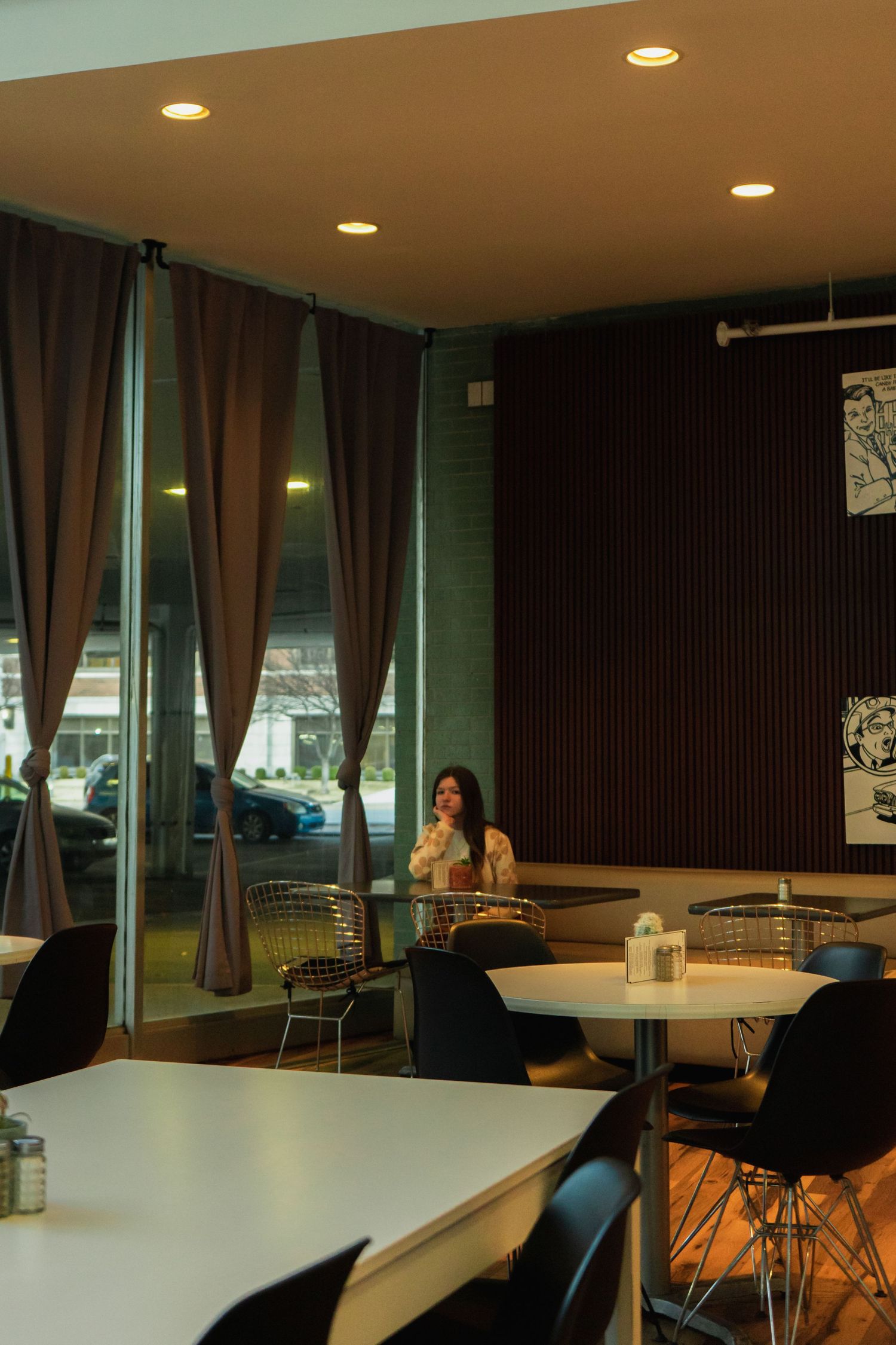 Modern cafe interior with brown curtains, white tables and black chairs under recessed lighting.