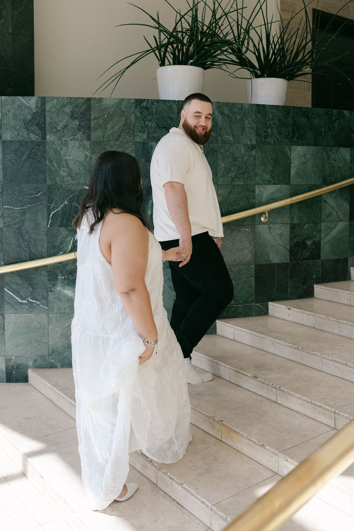 Couple in wedding attire walking up marble stairs, woman in white dress, man in cream shirt smiling back at camera.