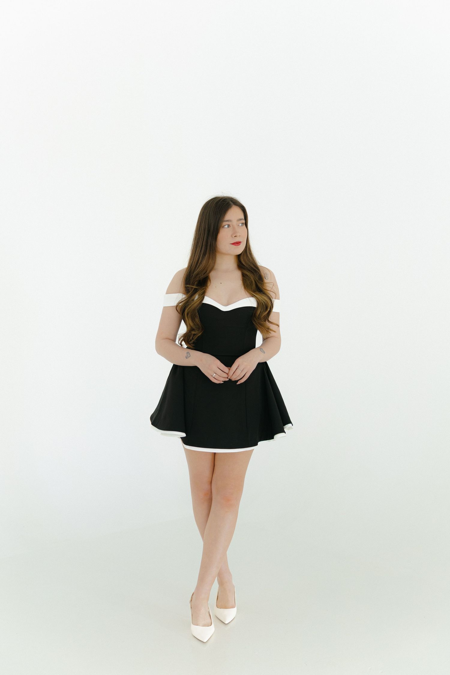 Young woman in black off-shoulder mini dress with white trim and heels poses against white background.