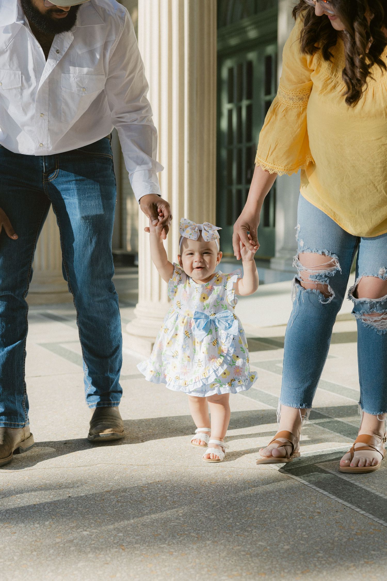 Small child in floral dress holding hands with parents while walking between columns on sunny day.