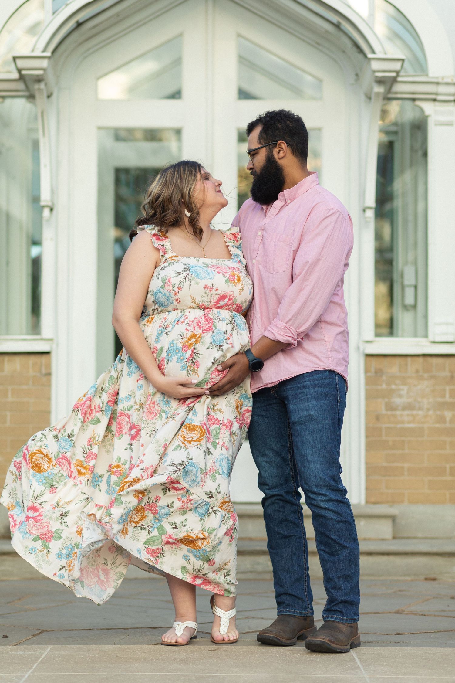 A couple in a romantic pose outdoors with a floral maternity dress against an architectural backdrop.