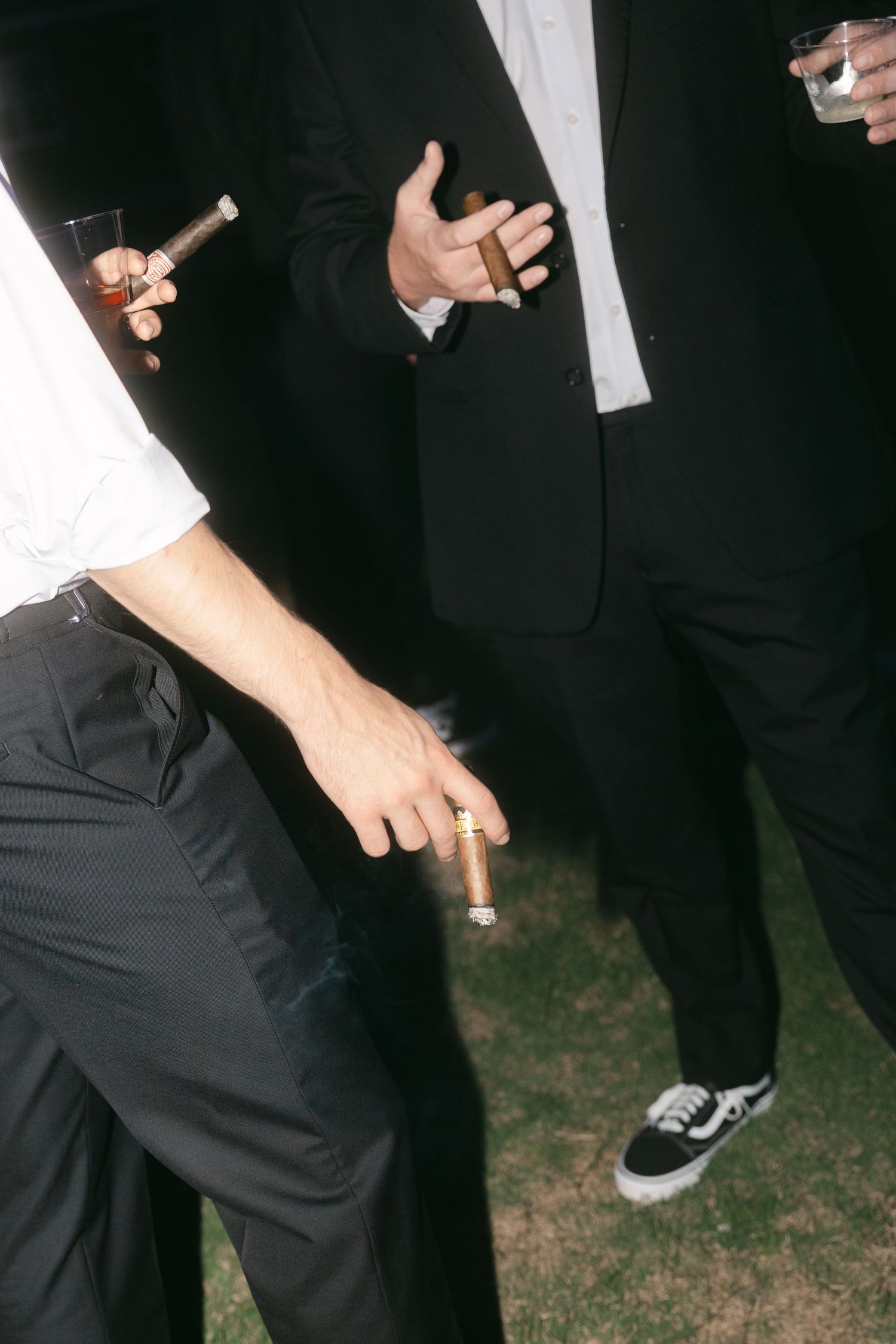 Men in formal suits holding cigars and drinks at an outdoor evening event.