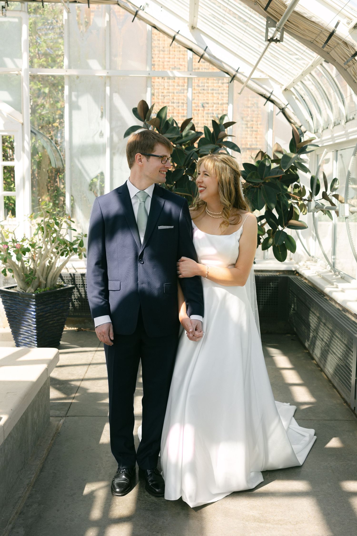 Bride and groom smiling at each other in a sunlit greenhouse, bride in white gown, groom in navy suit.