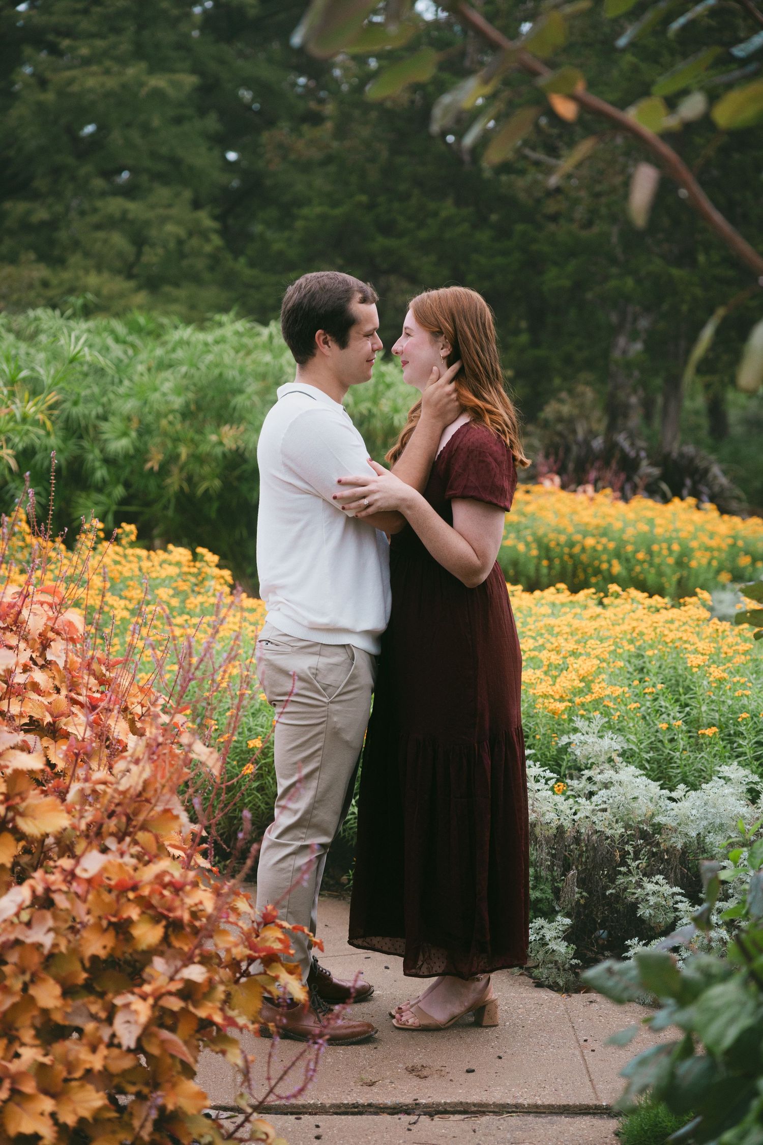 A couple embraces in a garden filled with colorful flowers, she in burgundy dress and he in white polo.