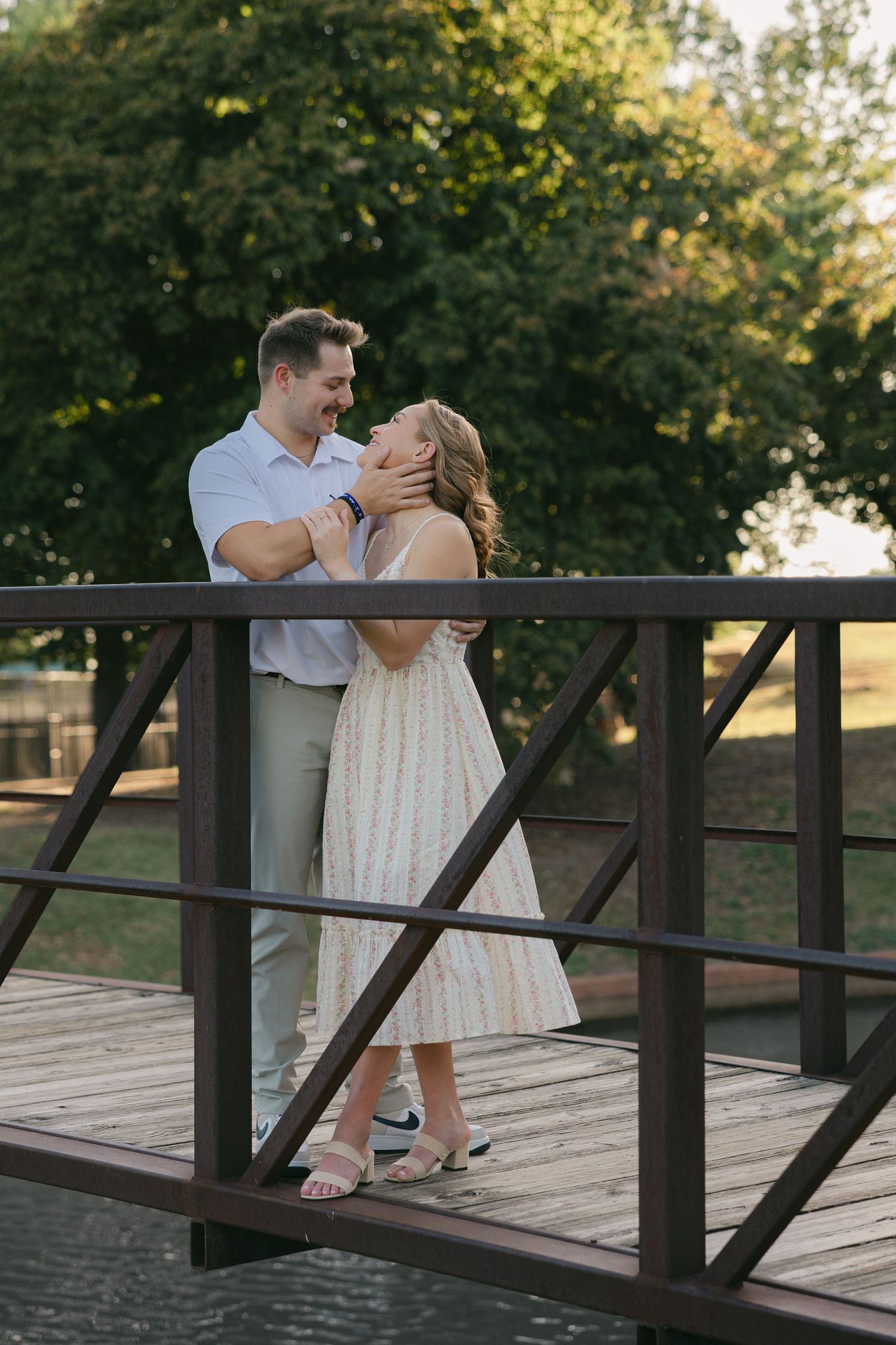 Couple sharing a romantic embrace on a wooden bridge surrounded by lush green trees during golden hour.