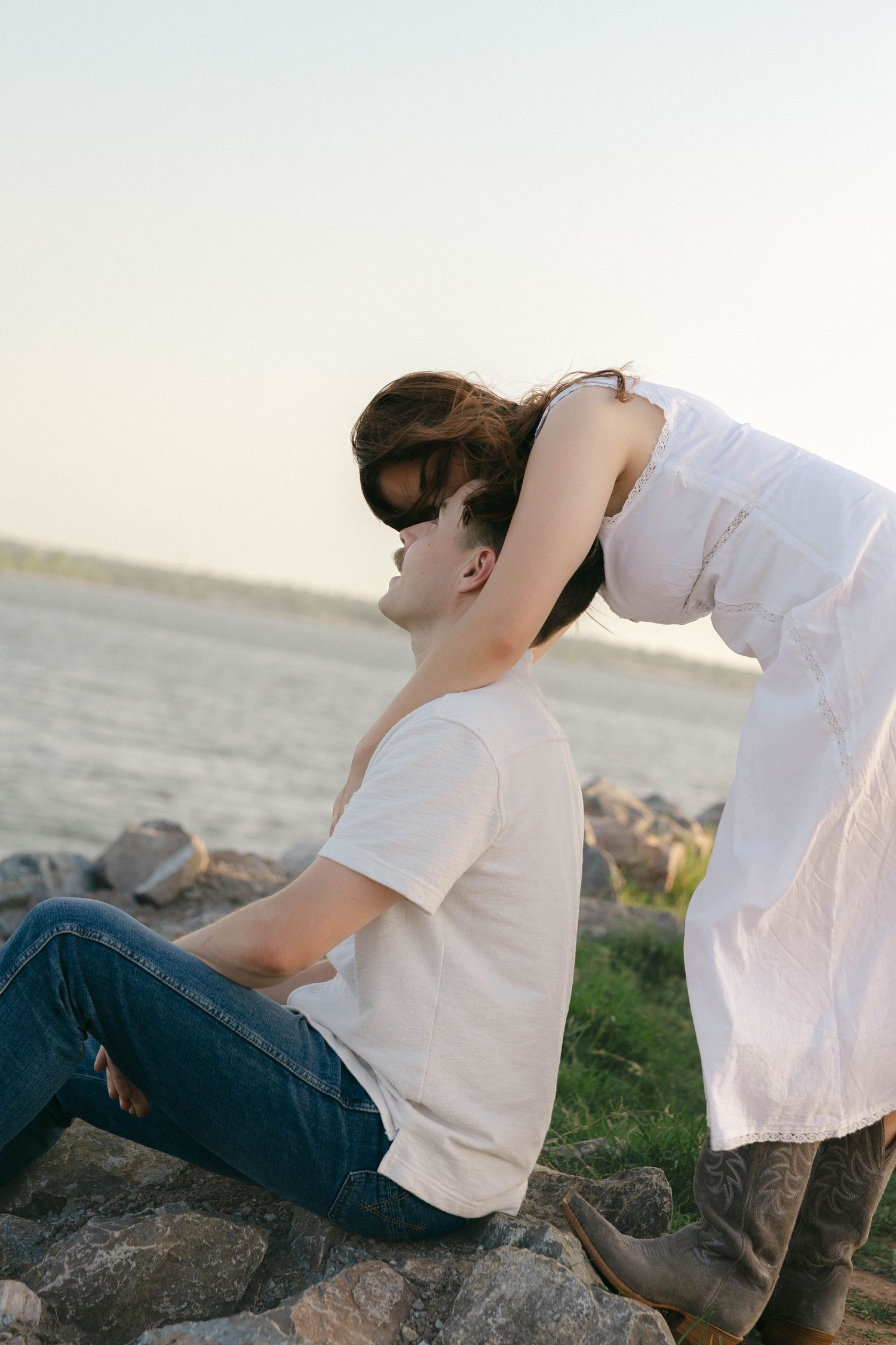 Couple sharing a tender moment on rocky shore, woman leaning over seated man near the water at sunset.