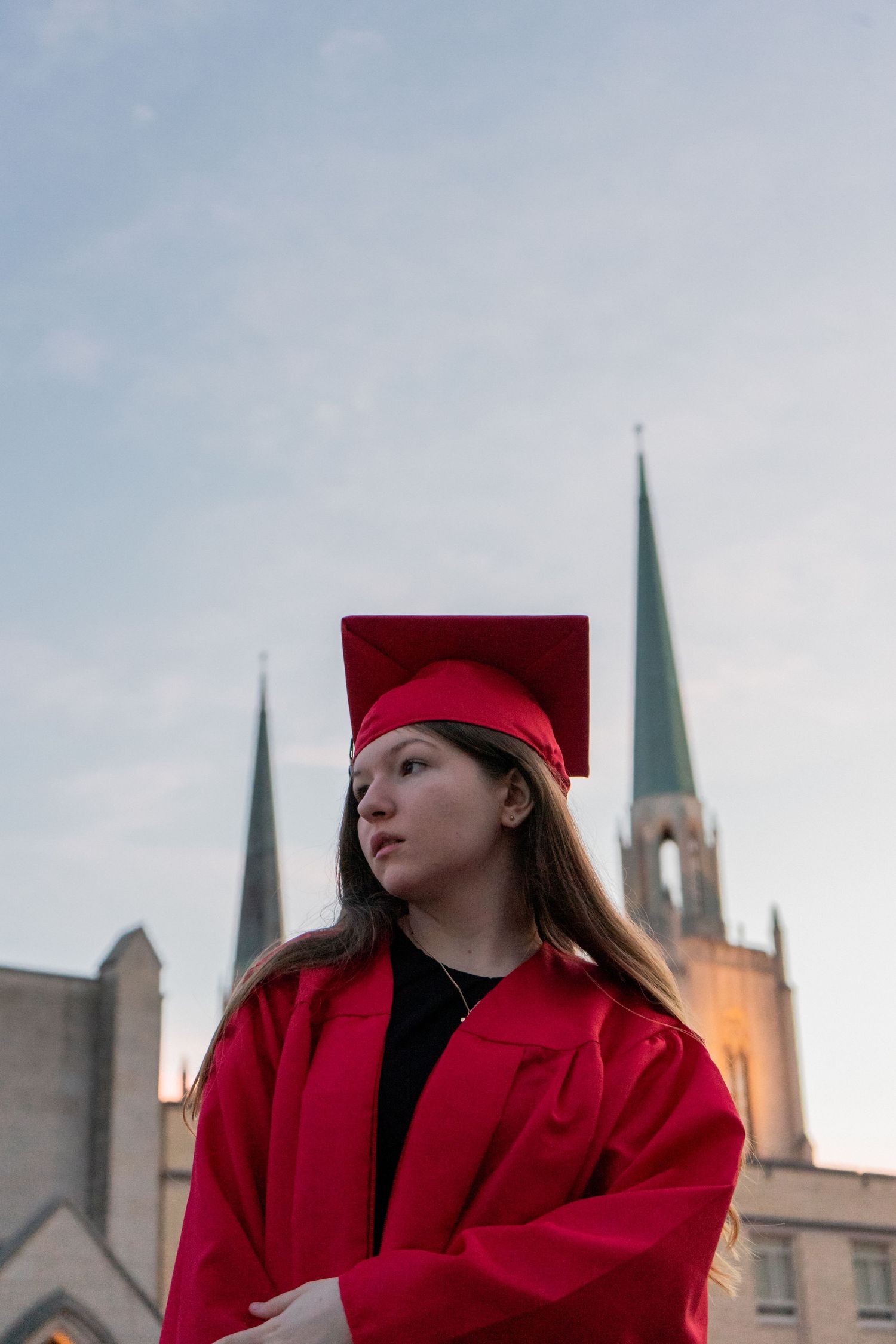 Graduate in bright red academic regalia poses dramatically with church spires visible in the background.