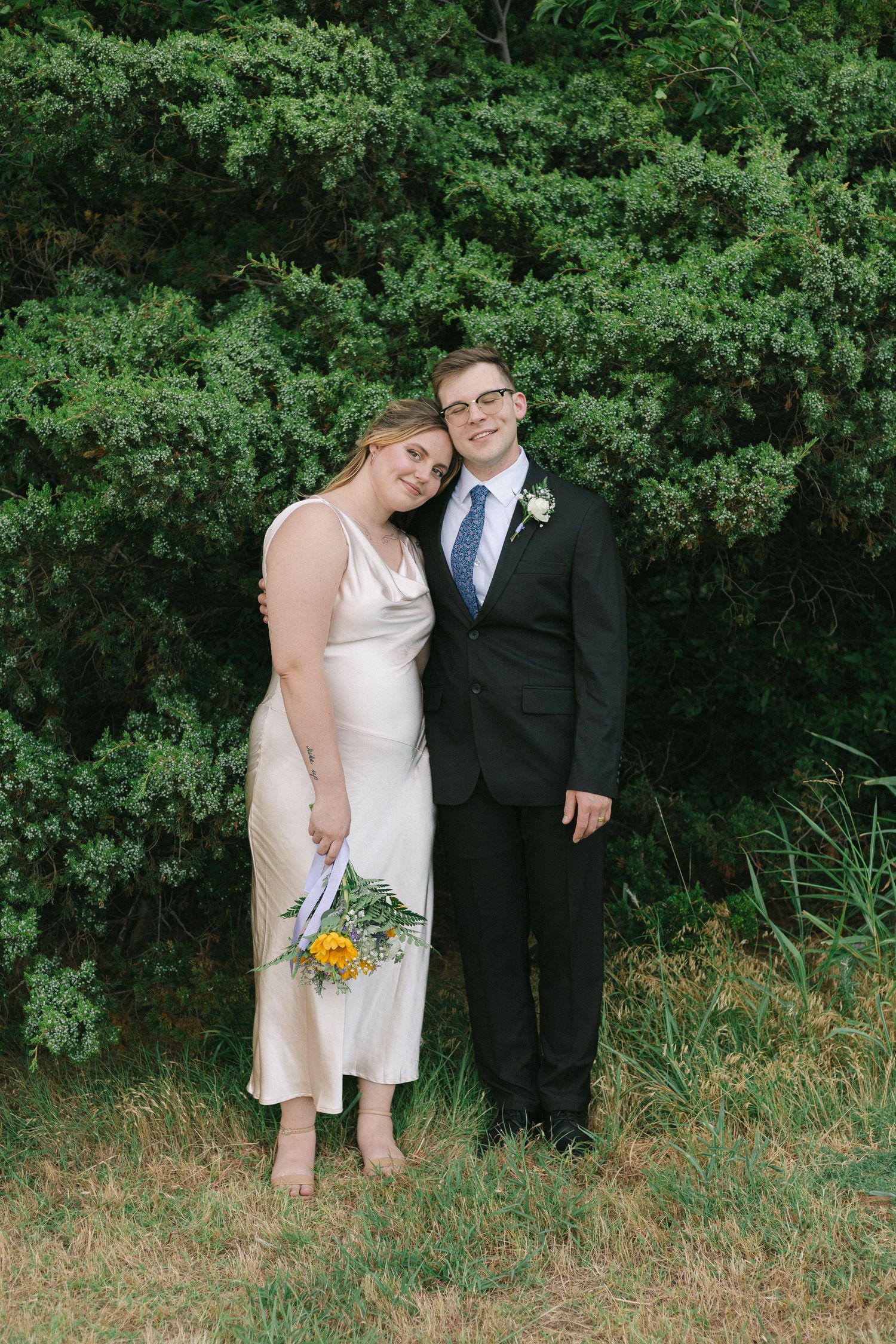 A joyful couple in wedding attire stands together against a backdrop of lush green foliage during their outdoor ceremony.