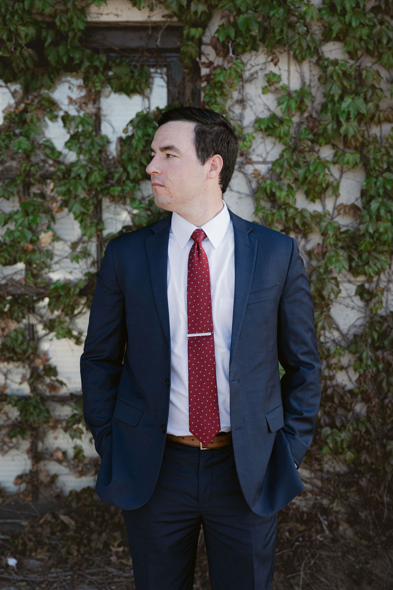 Professional in a navy suit and red tie stands against an ivy-covered wall in a series of poses.