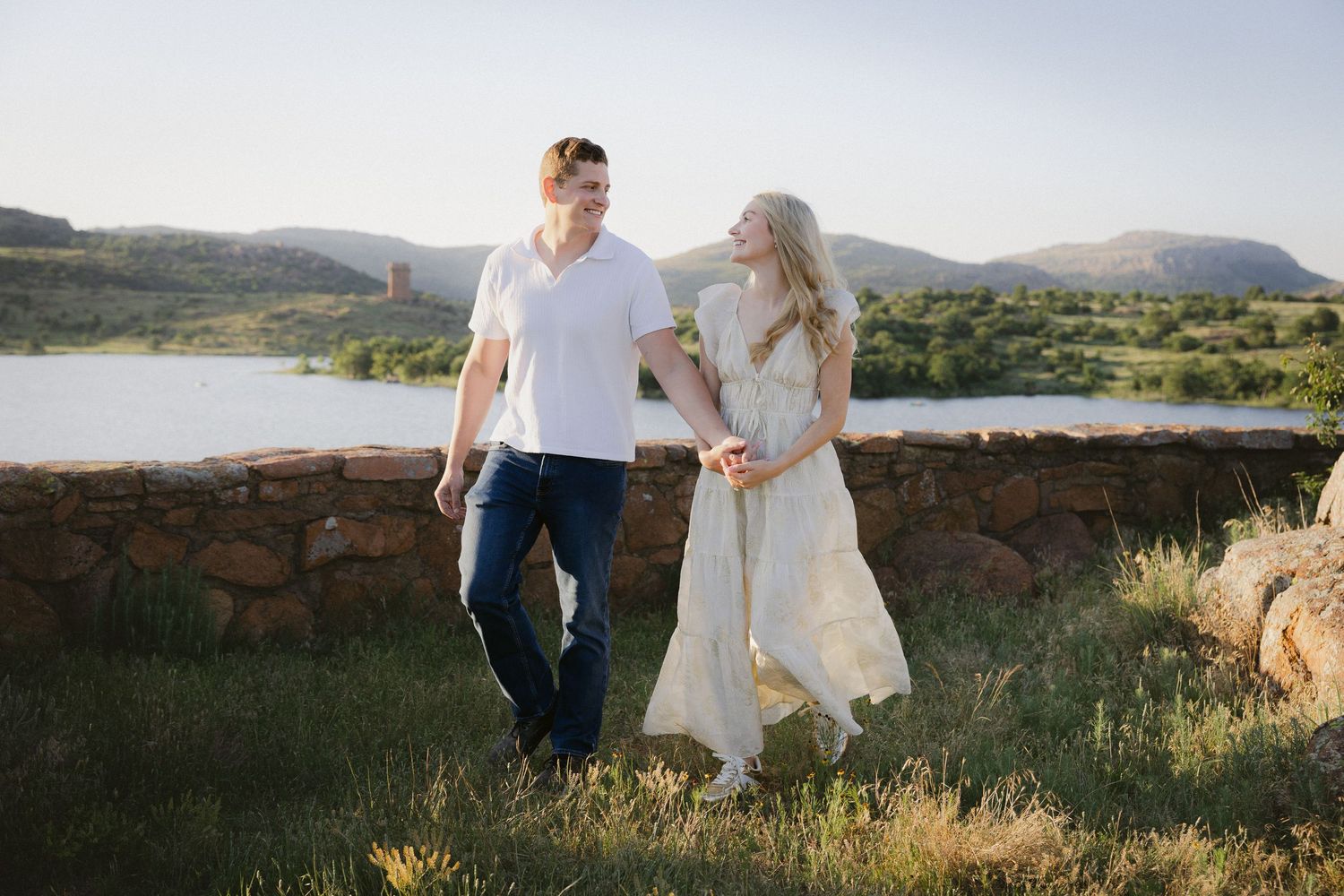A couple walks hand in hand through a sun-drenched mountain valley while wearing white and denim clothing.