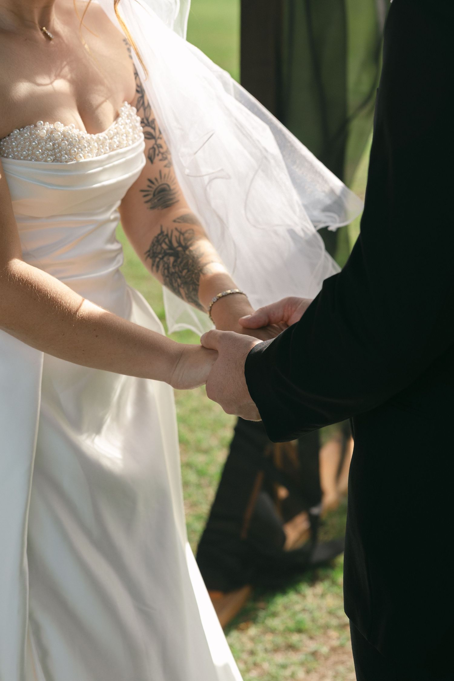 Bride with tattoos in white beaded gown holds grooms hands during outdoor wedding ceremony.