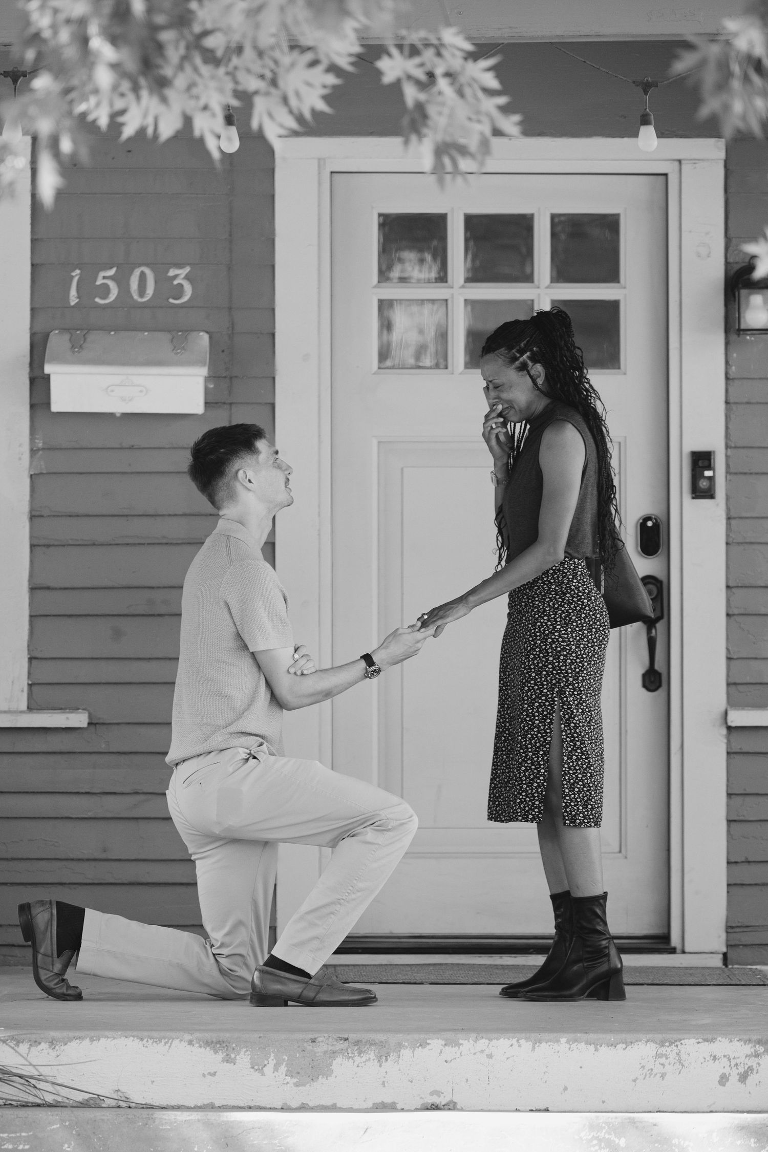 Man proposes on one knee to woman on front porch of house number 1503 in black and white photo.
