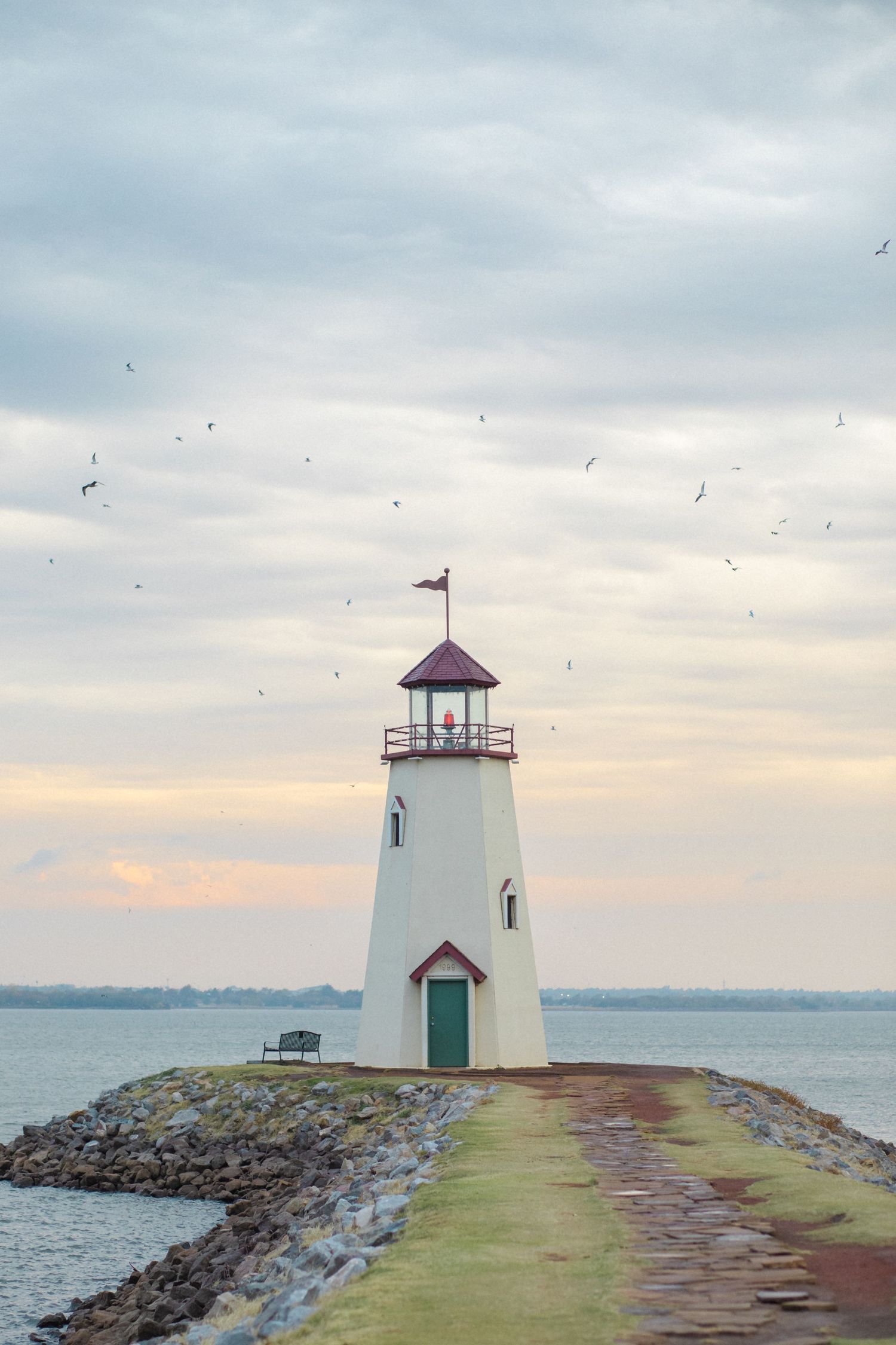 A small white lighthouse with a red roof stands at the end of a rocky breakwater during sunset over calm waters.