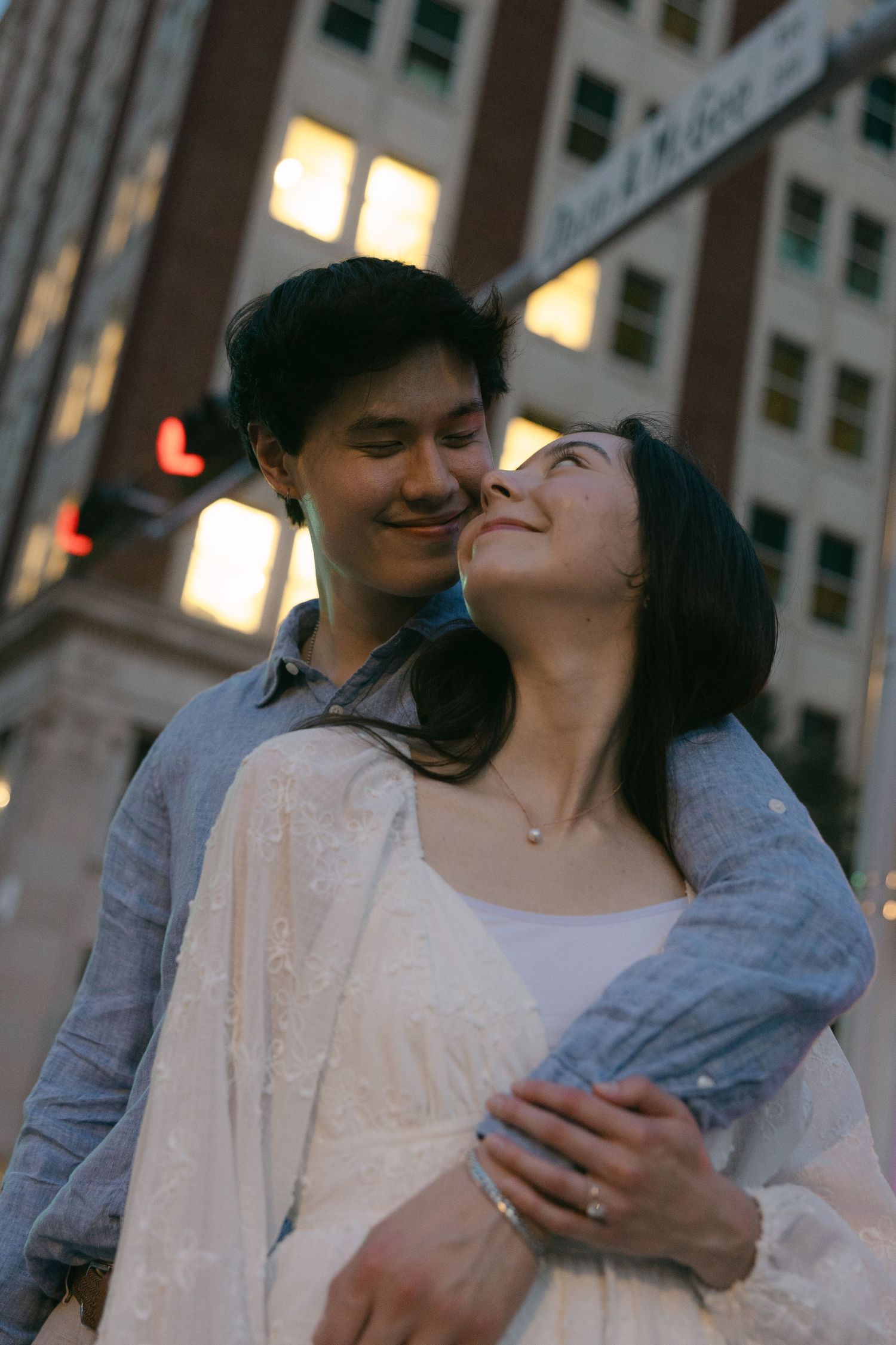 A young couple embraces and smiles in a city at dusk, with glowing building windows in the background.