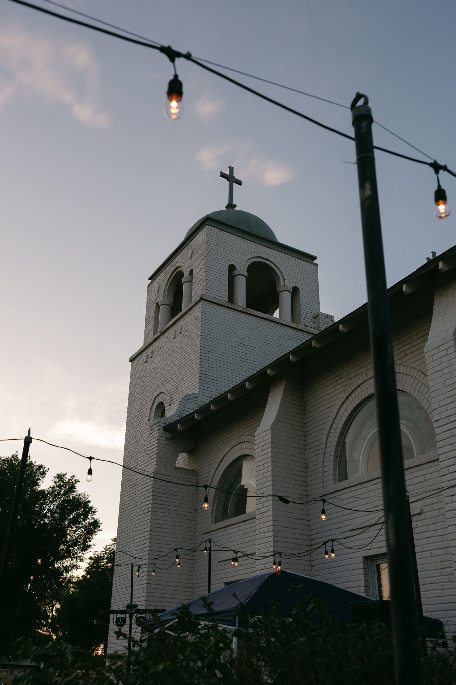 White brick church tower with cross at dusk, framed by string lights and a dark pole against a twilight sky.