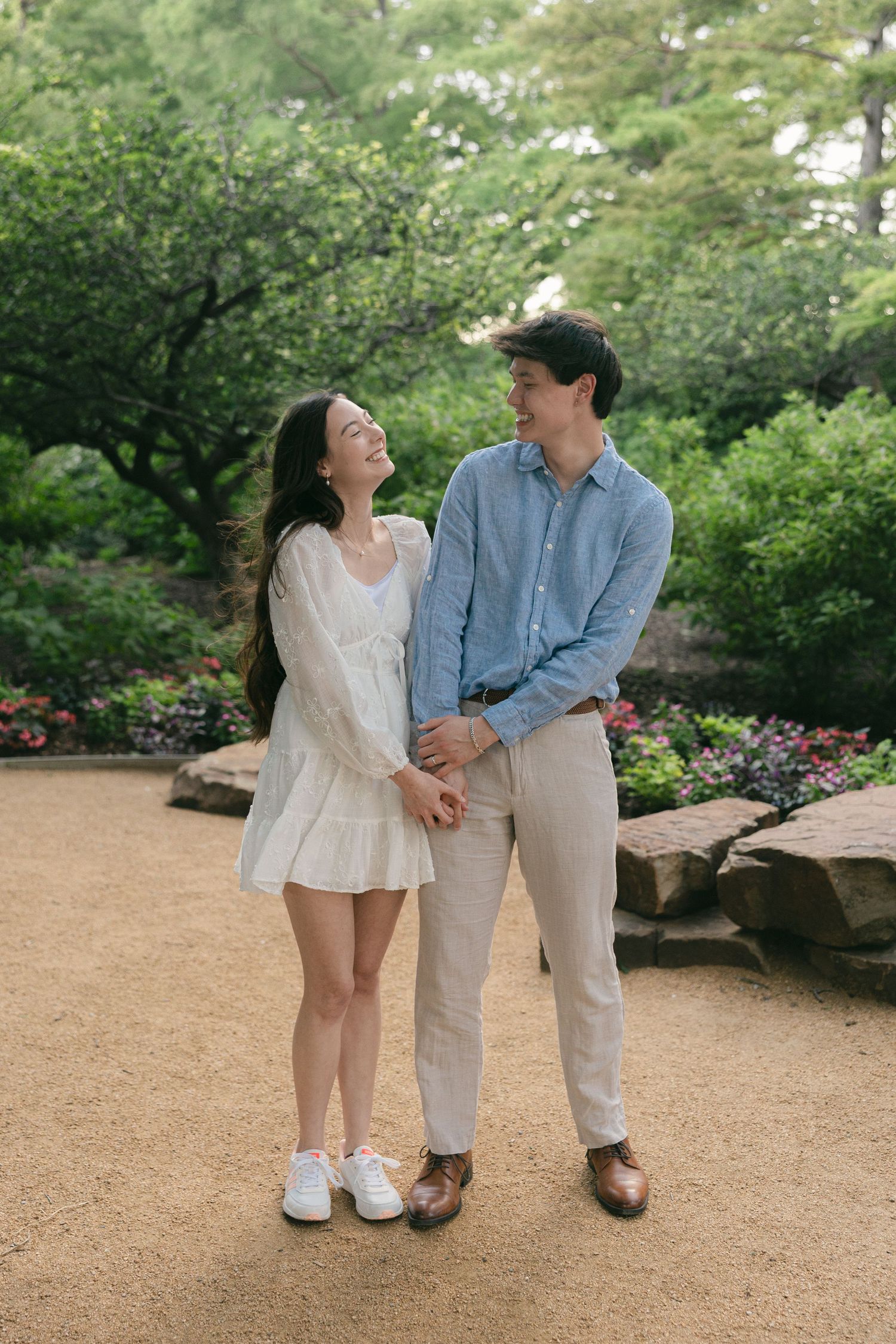 Couple holding hands and smiling at each other in a lush green garden, woman in white dress, man in blue shirt.