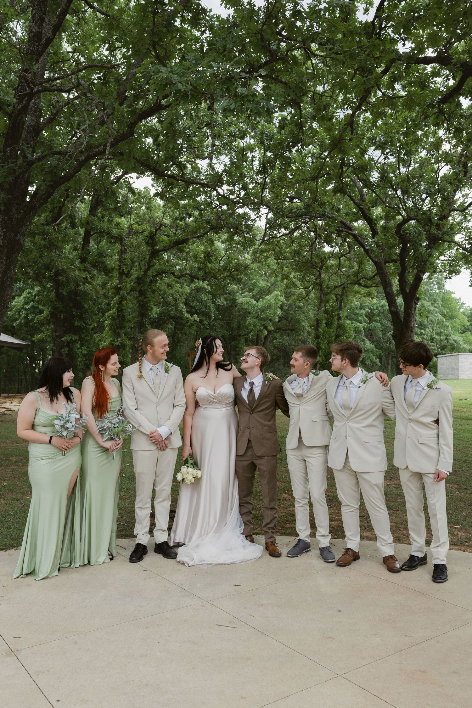 Wedding party poses together under trees wearing sage green dresses and light neutral suits.