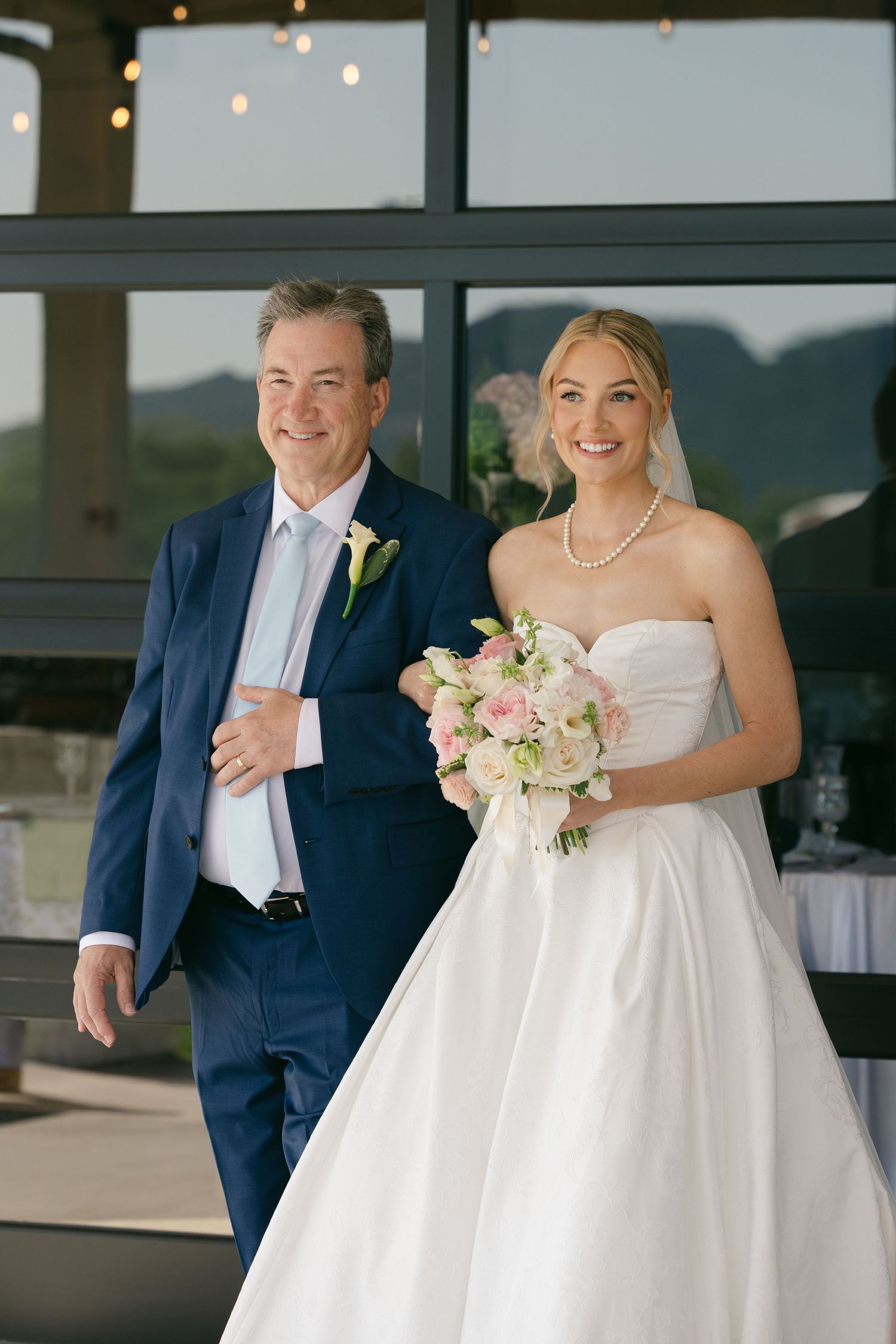 Smiling bride in strapless white gown holds pink bouquet while being escorted by man in navy suit.