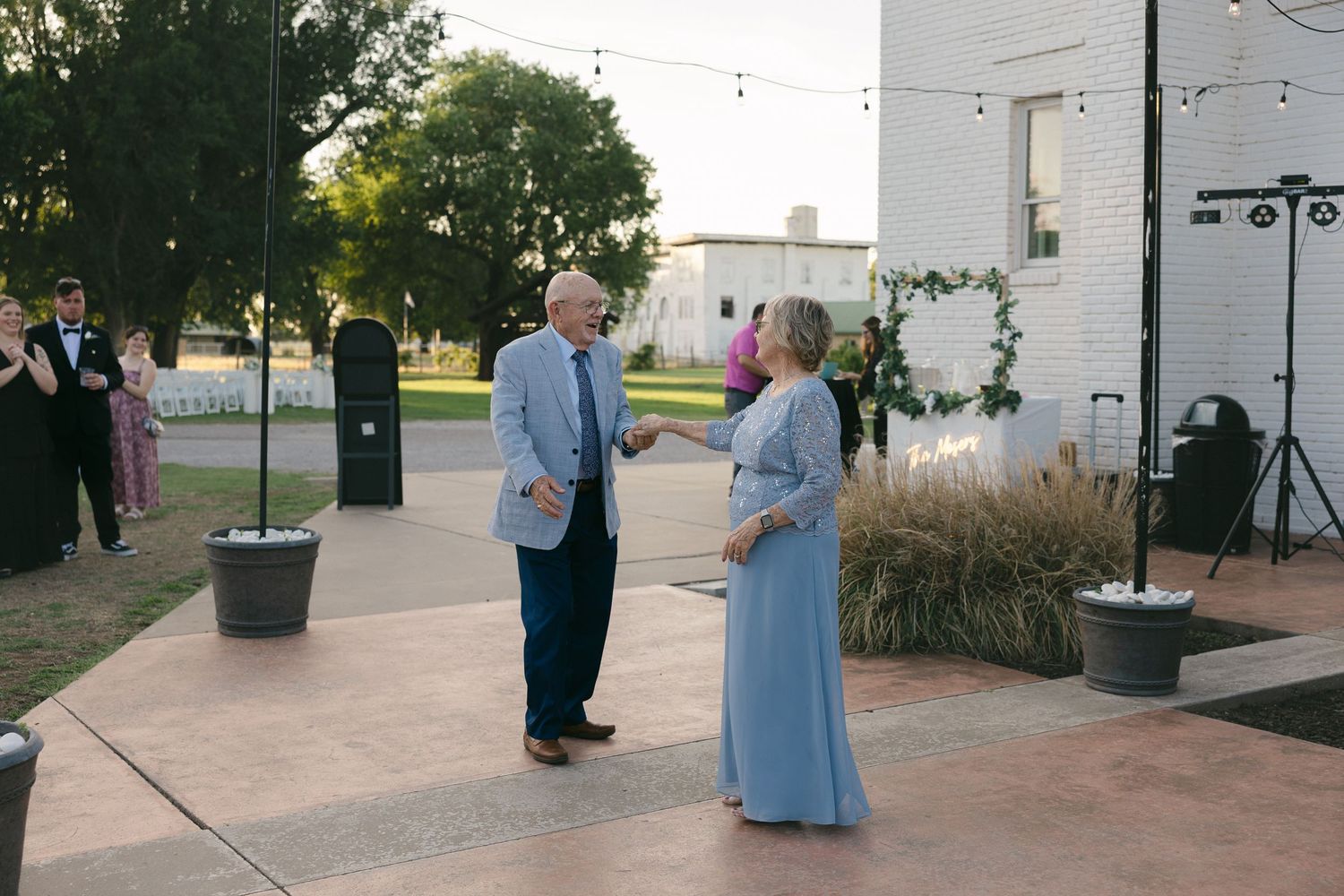 Elderly couple dancing outdoors at a wedding reception on a patio with string lights and white brick building.