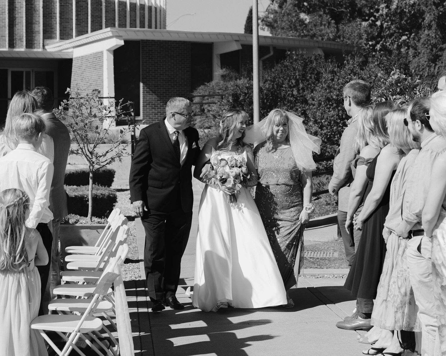 Black and white photo of a bride walking down the aisle with her father at an outdoor wedding ceremony.