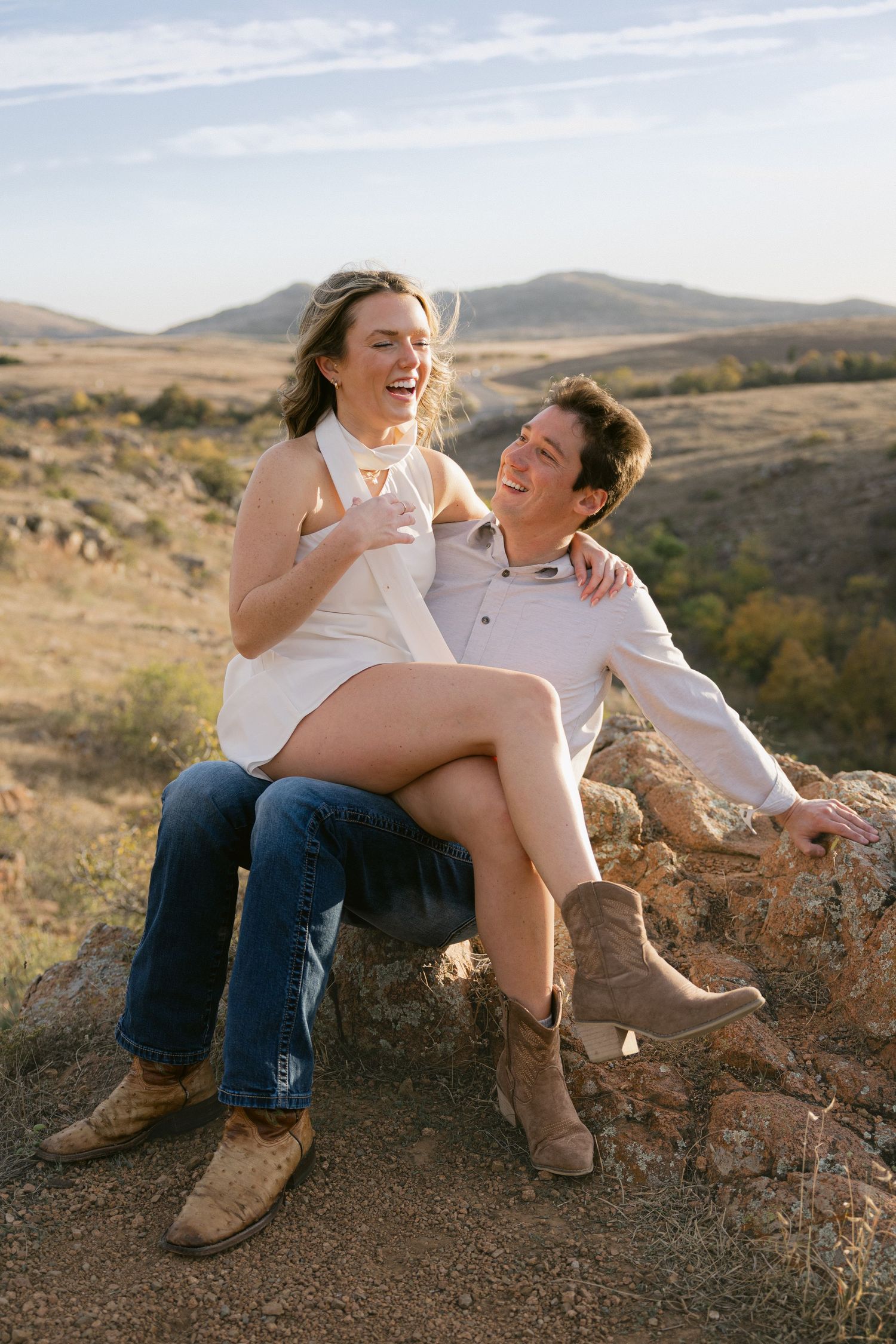 A couple shares a joyful moment while sitting together on rocks overlooking scenic desert hills at sunset.