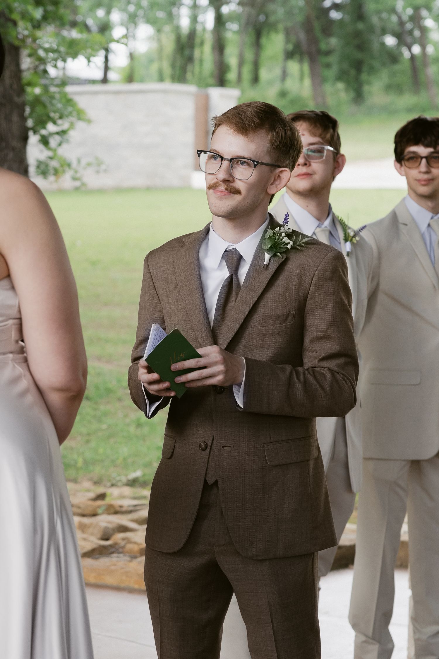 Groomsmen in formal suits stand attentively during an outdoor wedding ceremony.