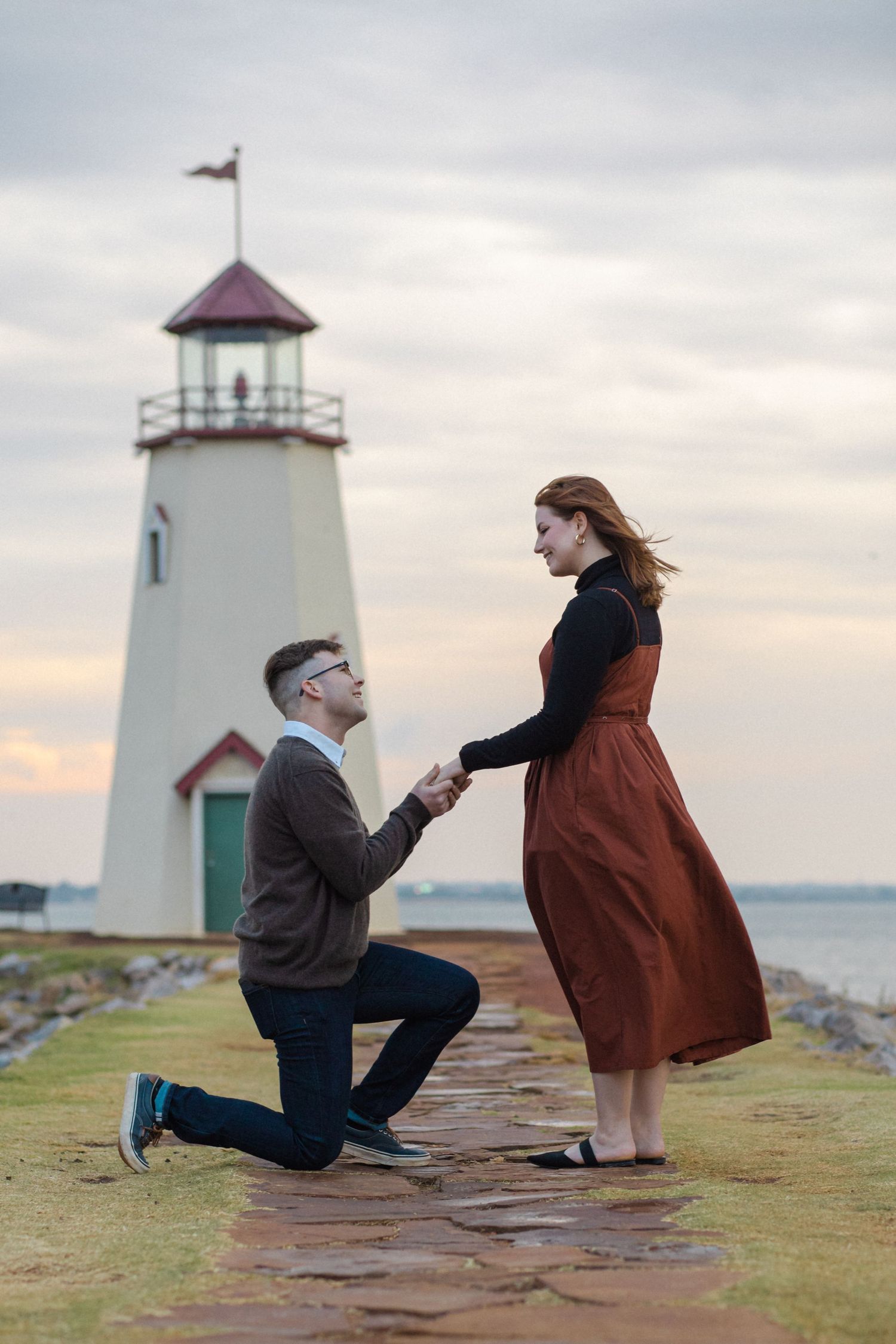 Scenic marriage proposal sequence on a coastal cliff near a lighthouse at sunset.