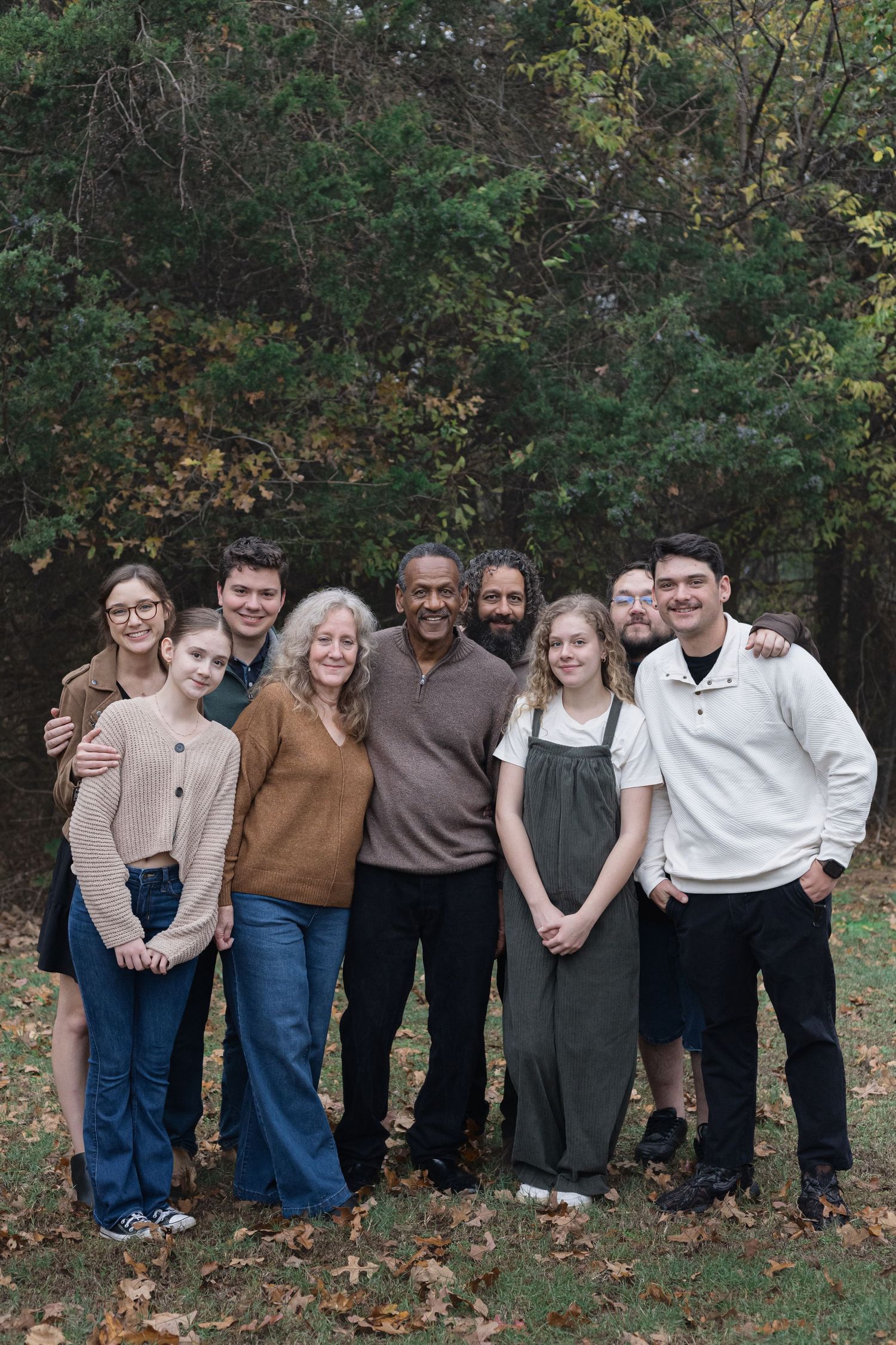 A group of people stand together outdoors in casual attire against a wooded backdrop during autumn.