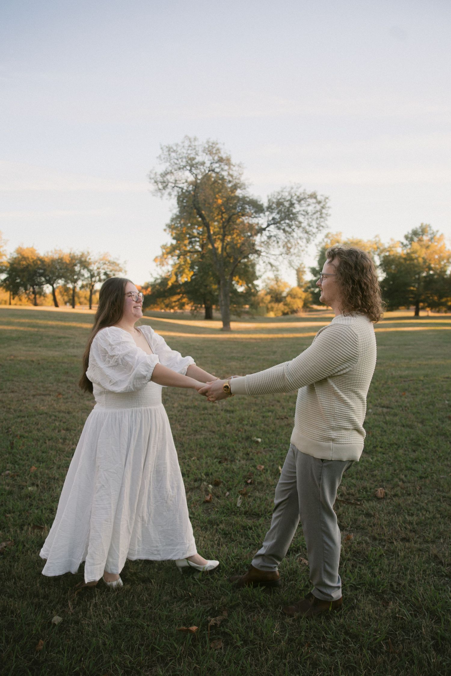 Two people hold hands and dance together in a park at sunset, one wearing a flowing white dress and the other in casual attire.