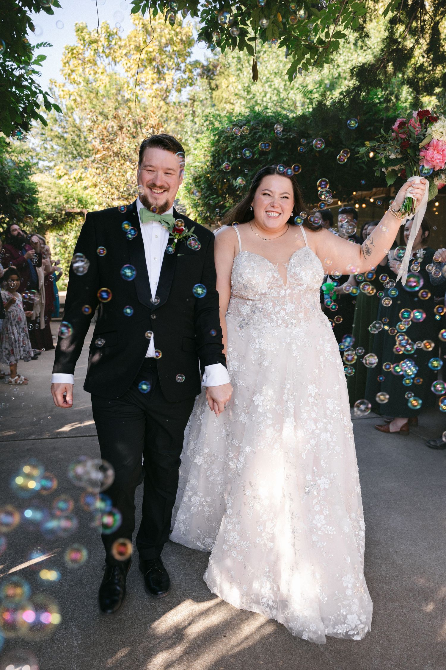 Wedding couple celebrates with joyful dance moves as soap bubbles float around them in an outdoor garden setting.