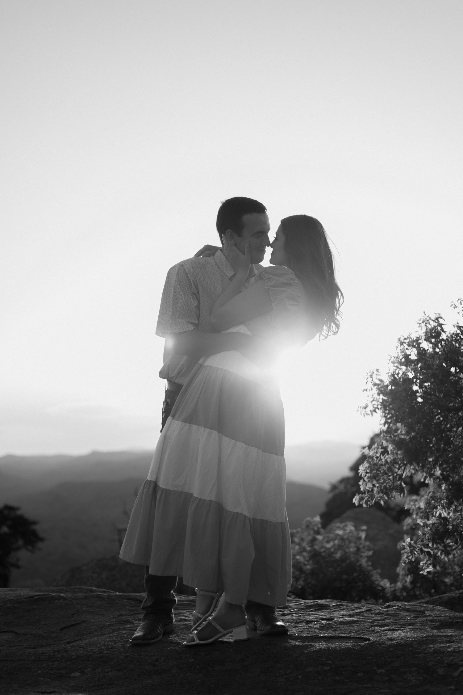 Romantic silhouetted figures embrace at sunset on a hilltop with dramatic backlighting and mountain views in the background.
