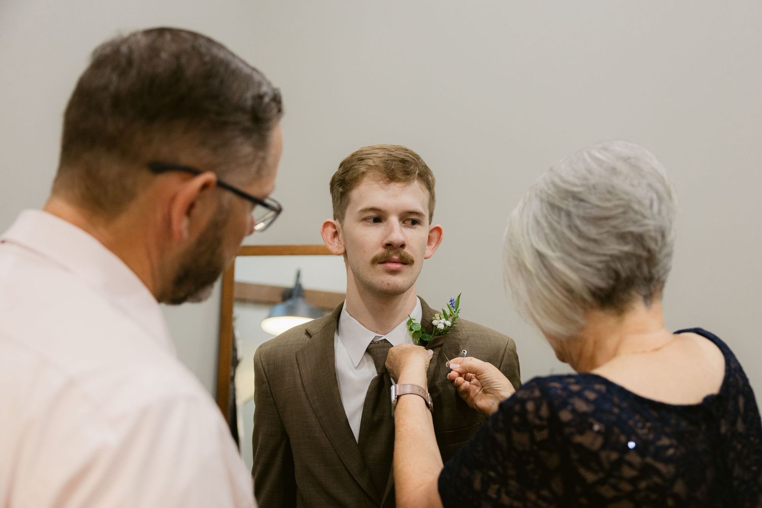 Someone receives a boutonniere pinned to their suit jacket while wearing a brown suit and tie at a formal event.