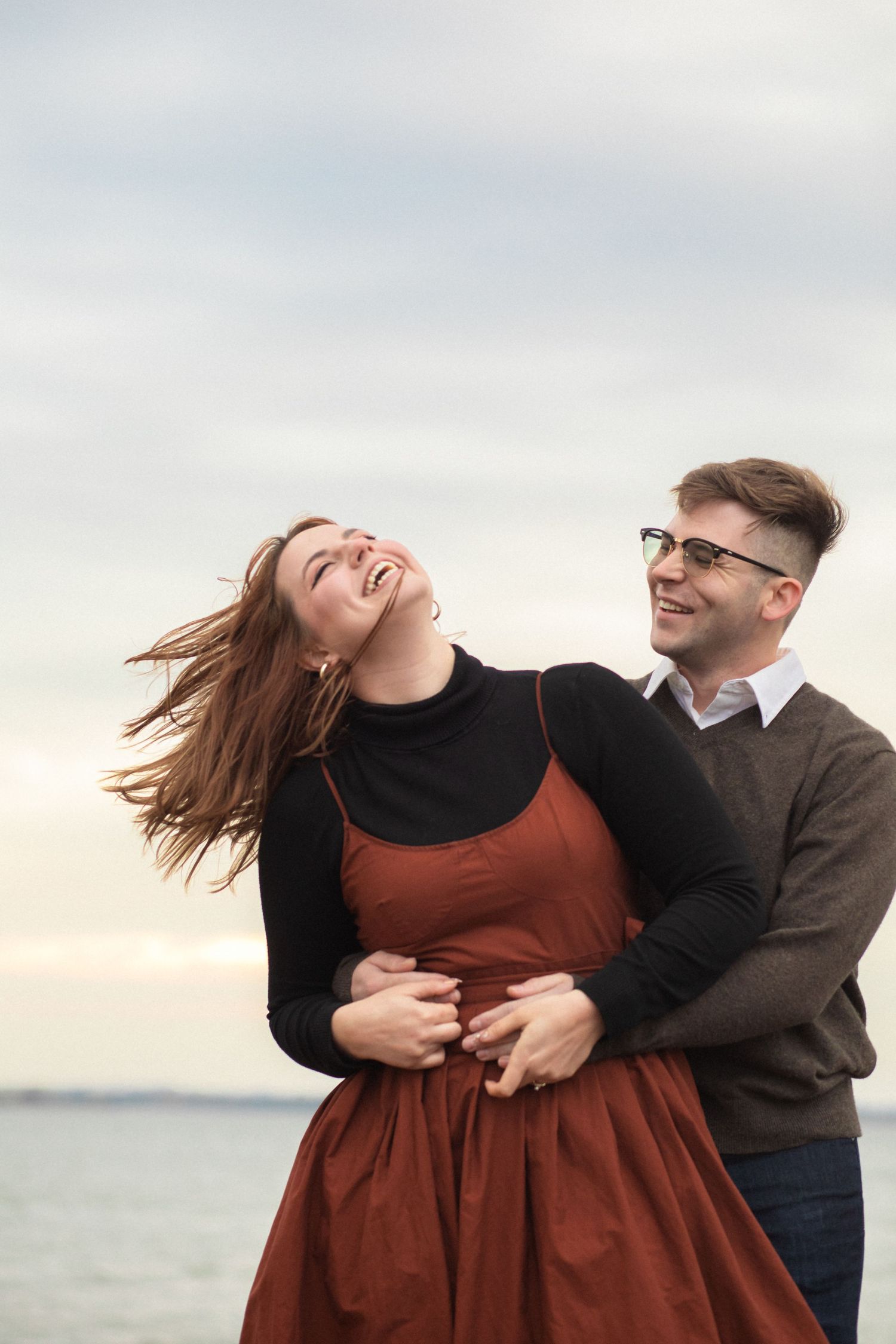 A playful couple laughs and embraces while posing for photos by the ocean during golden hour.