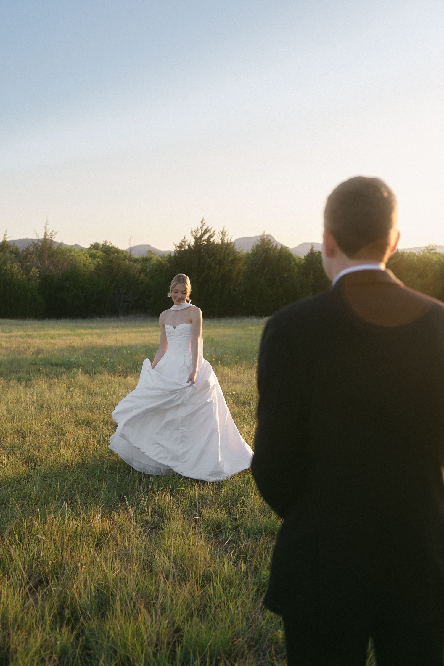 Bride in white gown walks toward groom in black tuxedo across sunlit green field at golden hour.