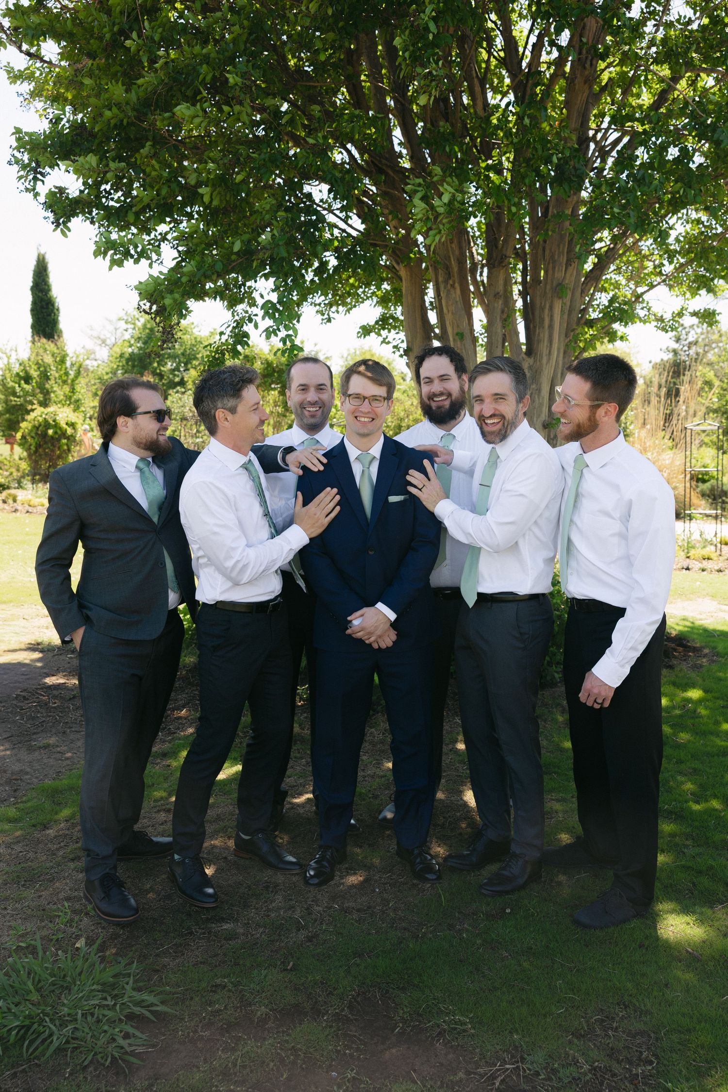 Groom surrounded by smiling groomsmen in mint ties and dark suits, laughing together outdoors under a tree.