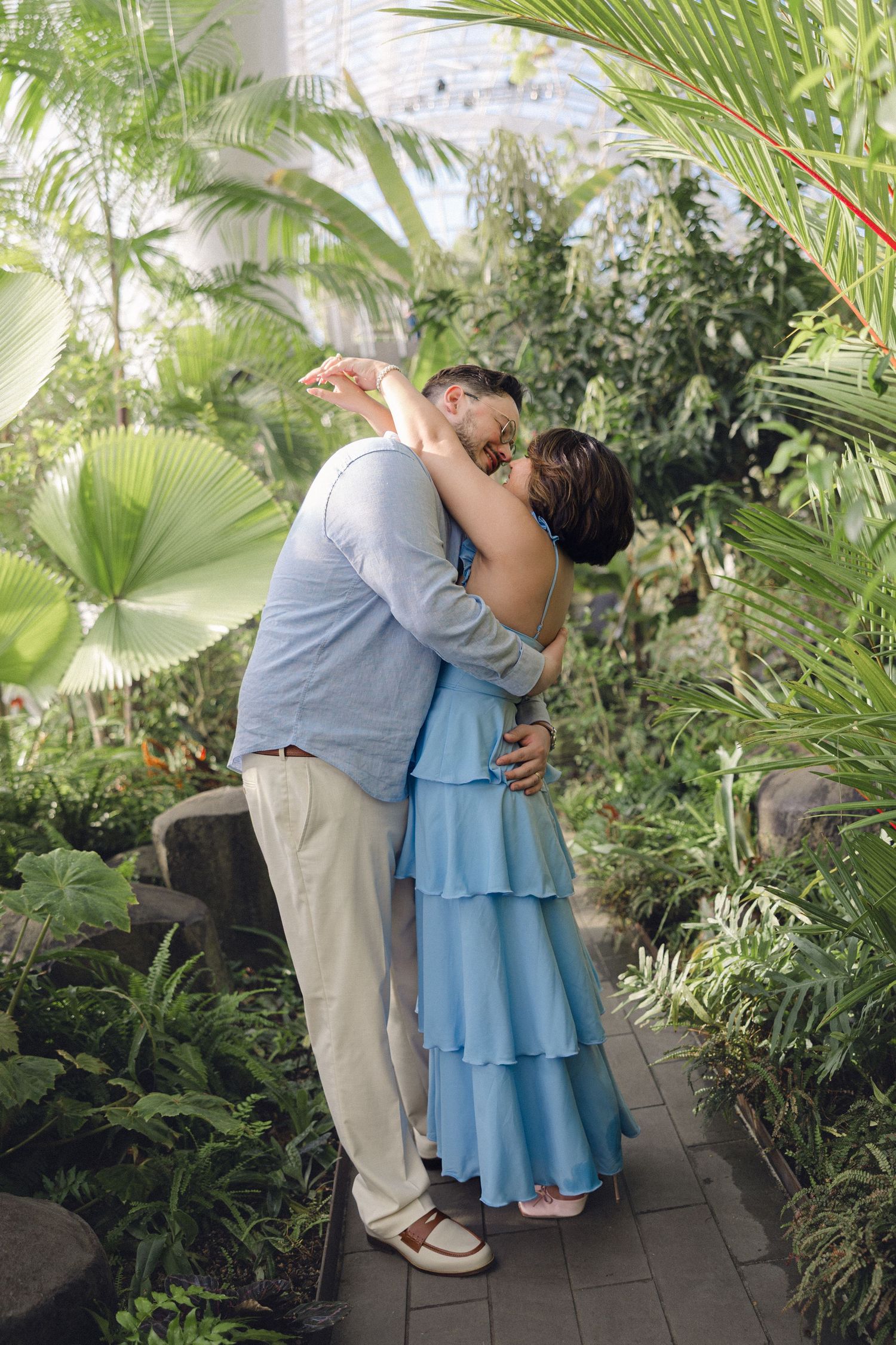 Couple sharing a romantic kiss in a lush tropical garden, woman in blue ruffled dress embracing man in casual attire.