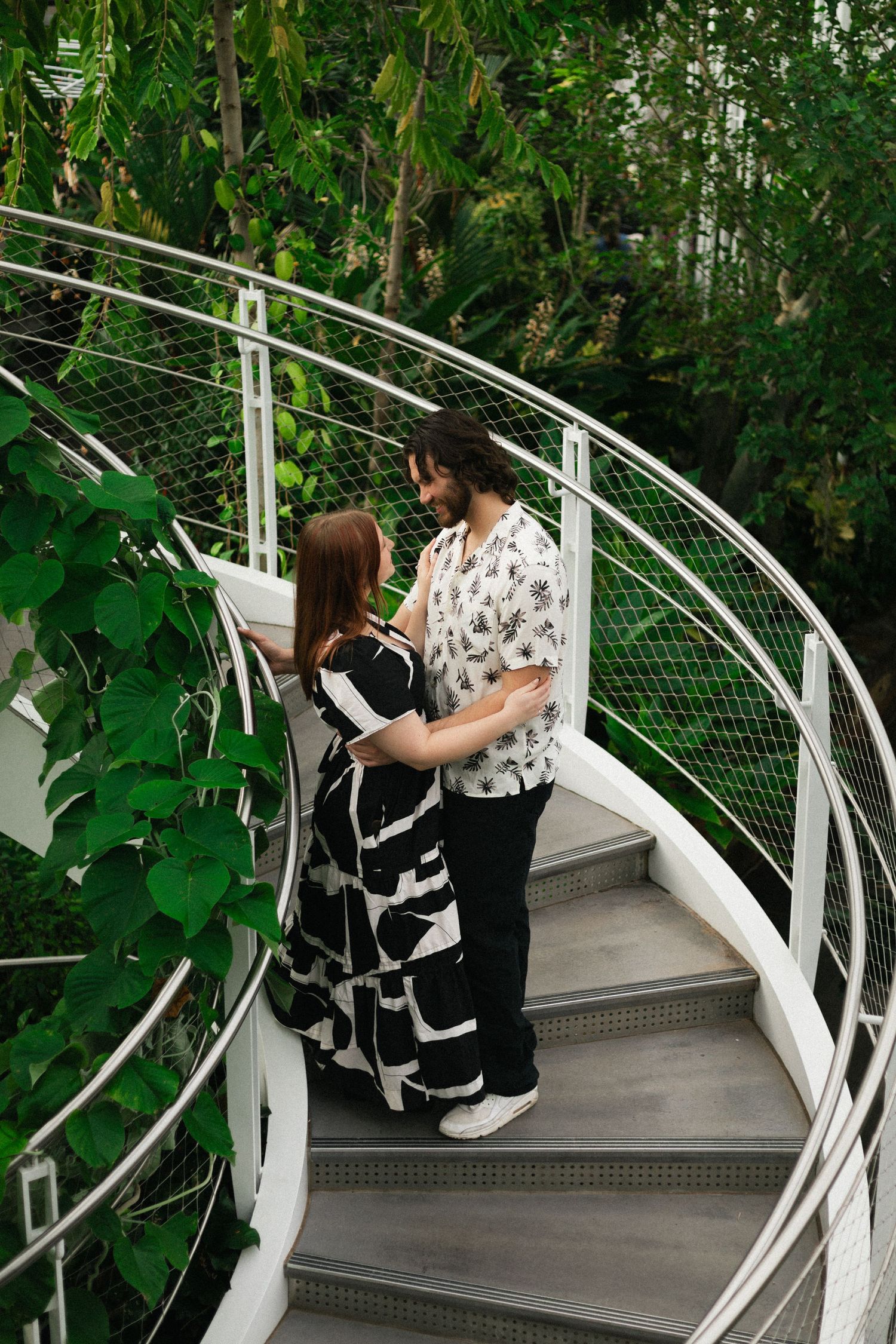 A couple embraces on a curved white staircase surrounded by lush greenery.