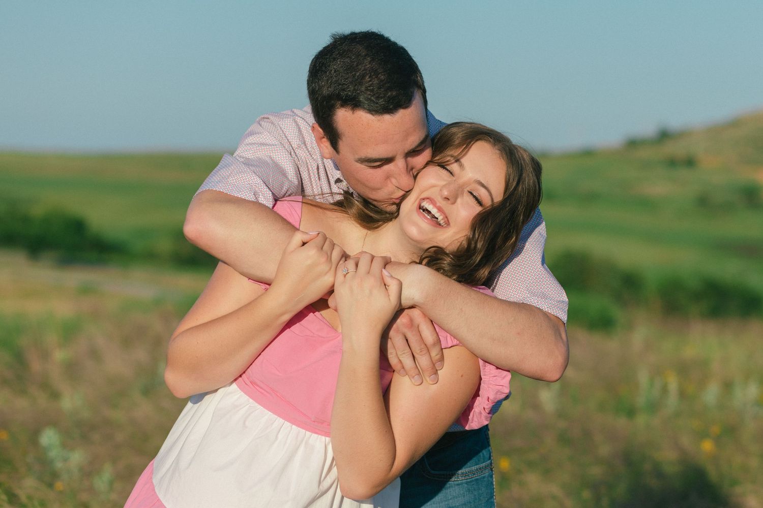 A joyful couple embraces in a sunny meadow with rolling green hills in the background.