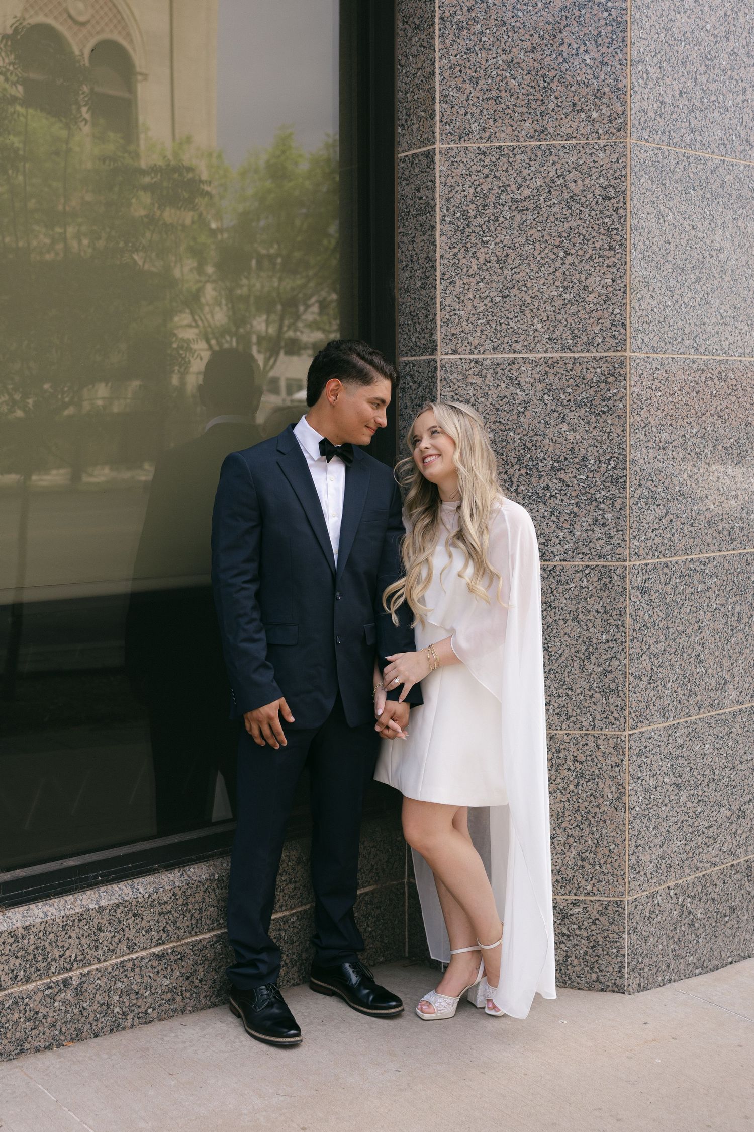 Elegant couple in wedding attire posing against a granite wall, groom in navy suit and bride in white mini dress.