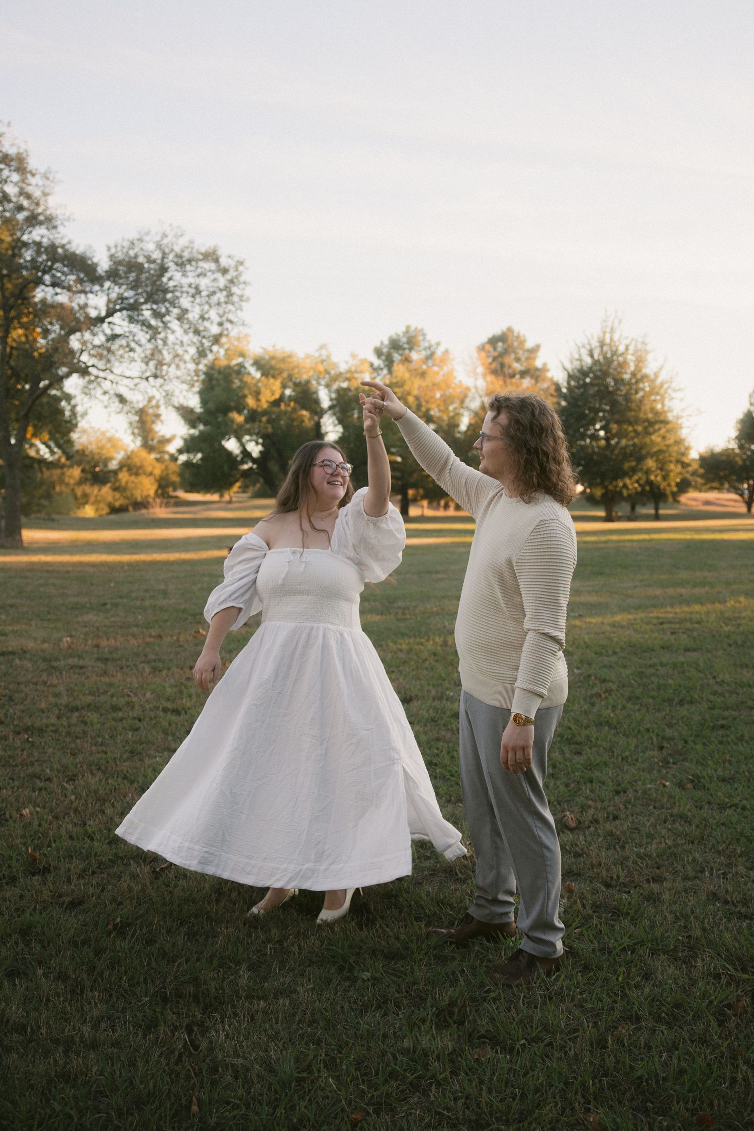 A couple in white and gray attire dances joyfully together in a grassy field at sunset.