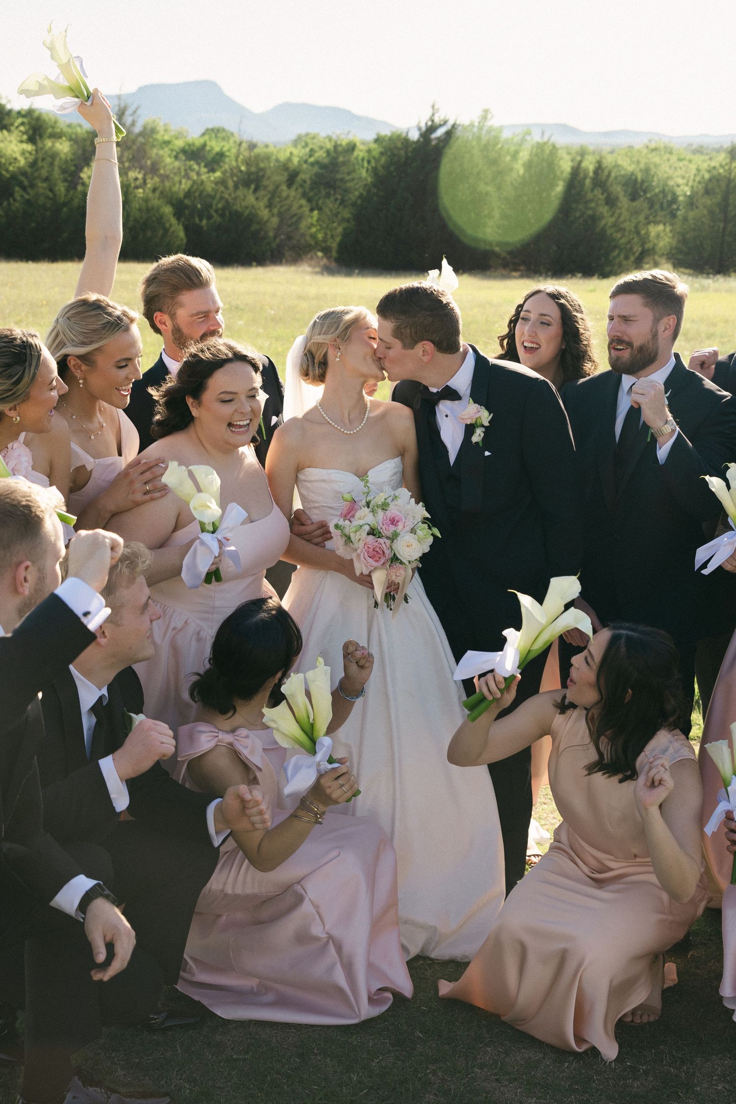 Bride and groom share a kiss surrounded by cheering wedding party in blush pink bridesmaid dresses outdoors.