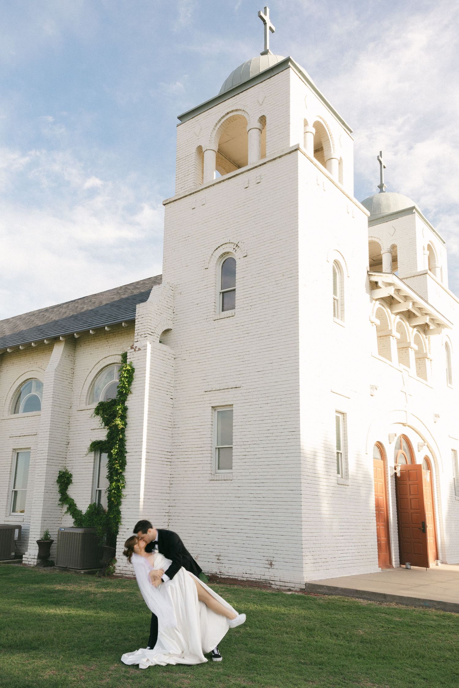Groom lifts bride in wedding gown outside a stunning white brick church with a tall bell tower.