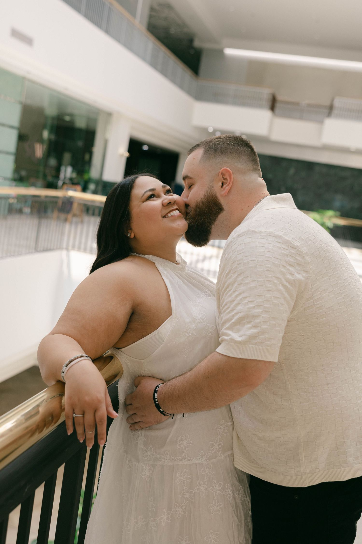 Couple in white attire sharing a romantic moment on a balcony, woman smiling as man kisses her cheek.