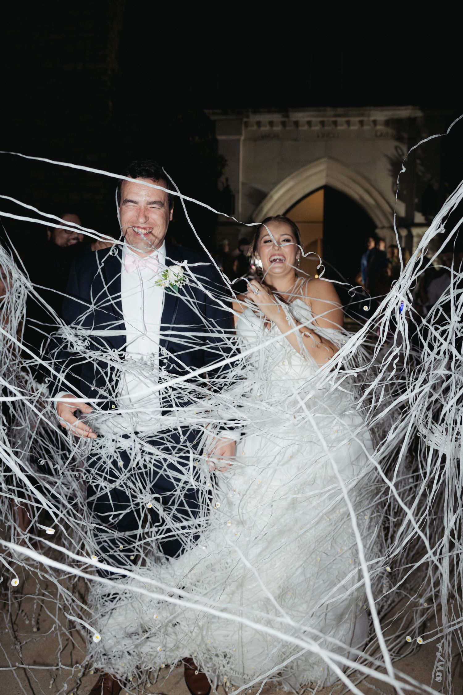Wedding couple bursting through decorative white streamers at night celebration.