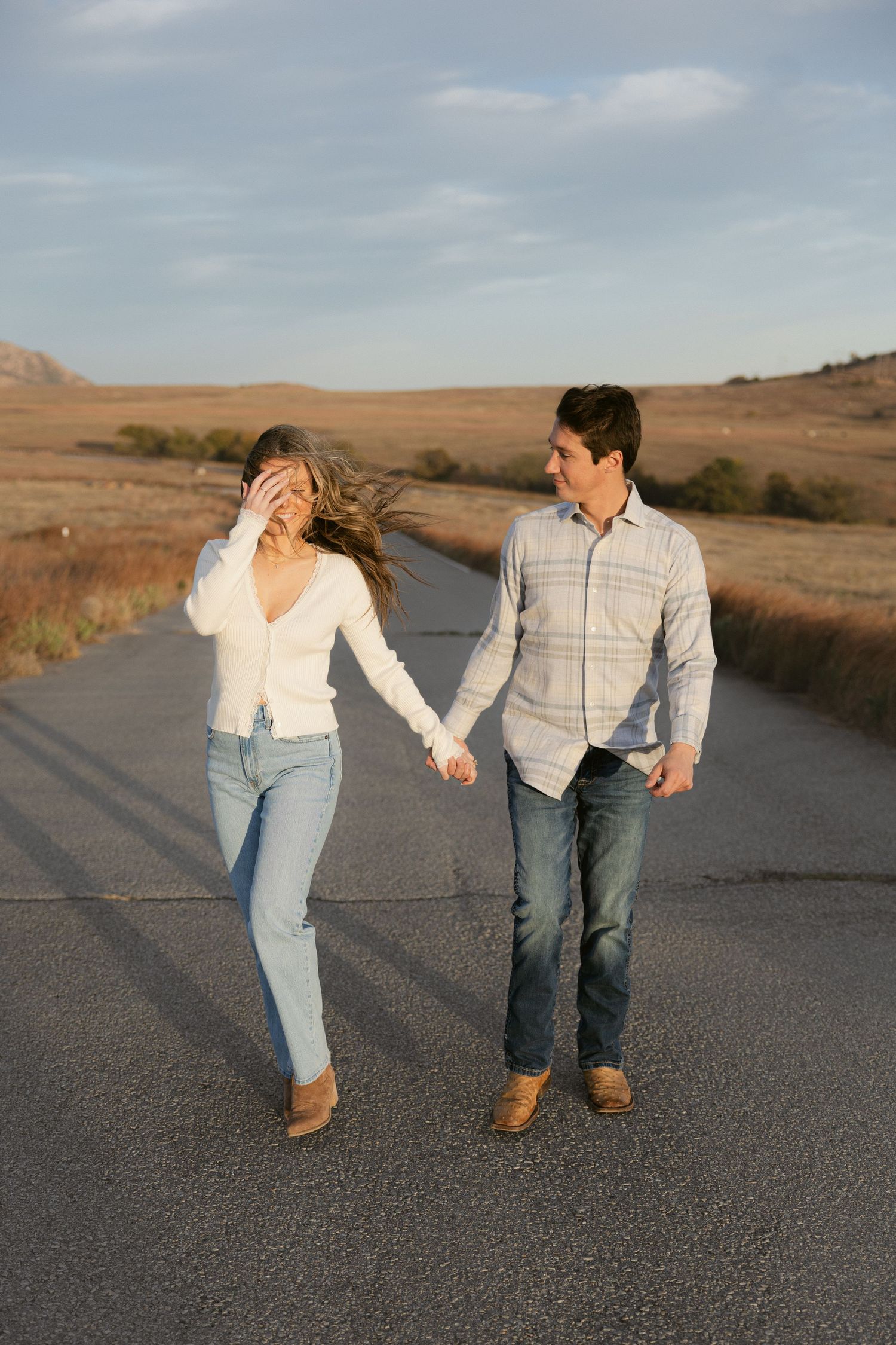 A couple holds hands and dances playfully on an empty desert road at sunset.