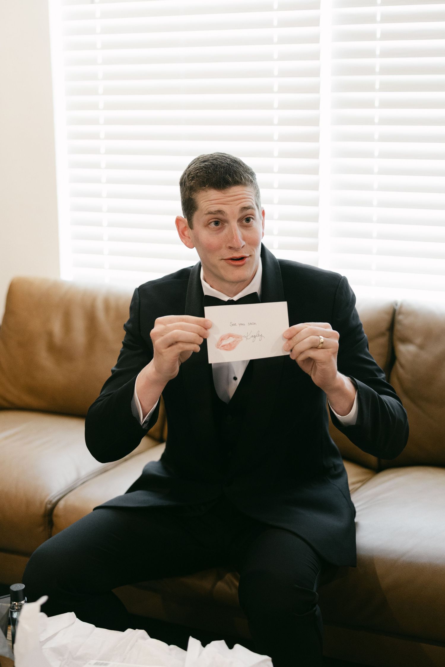 Groom in black tuxedo sitting on leather sofa holding a card with a lipstick kiss mark on his wedding day.