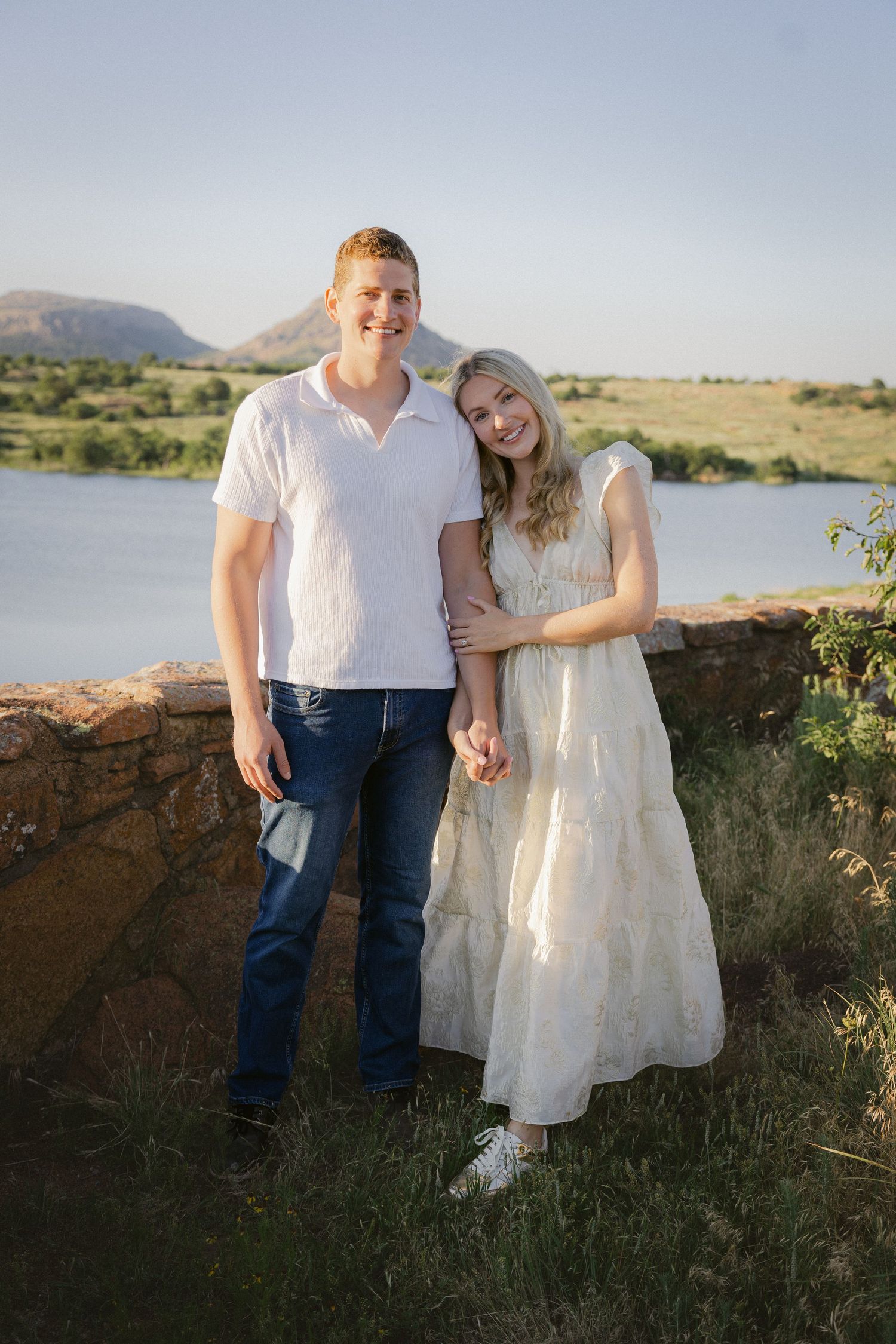 A couple embraces while standing together outdoors against a scenic backdrop of rolling hills and a sunlit lake.